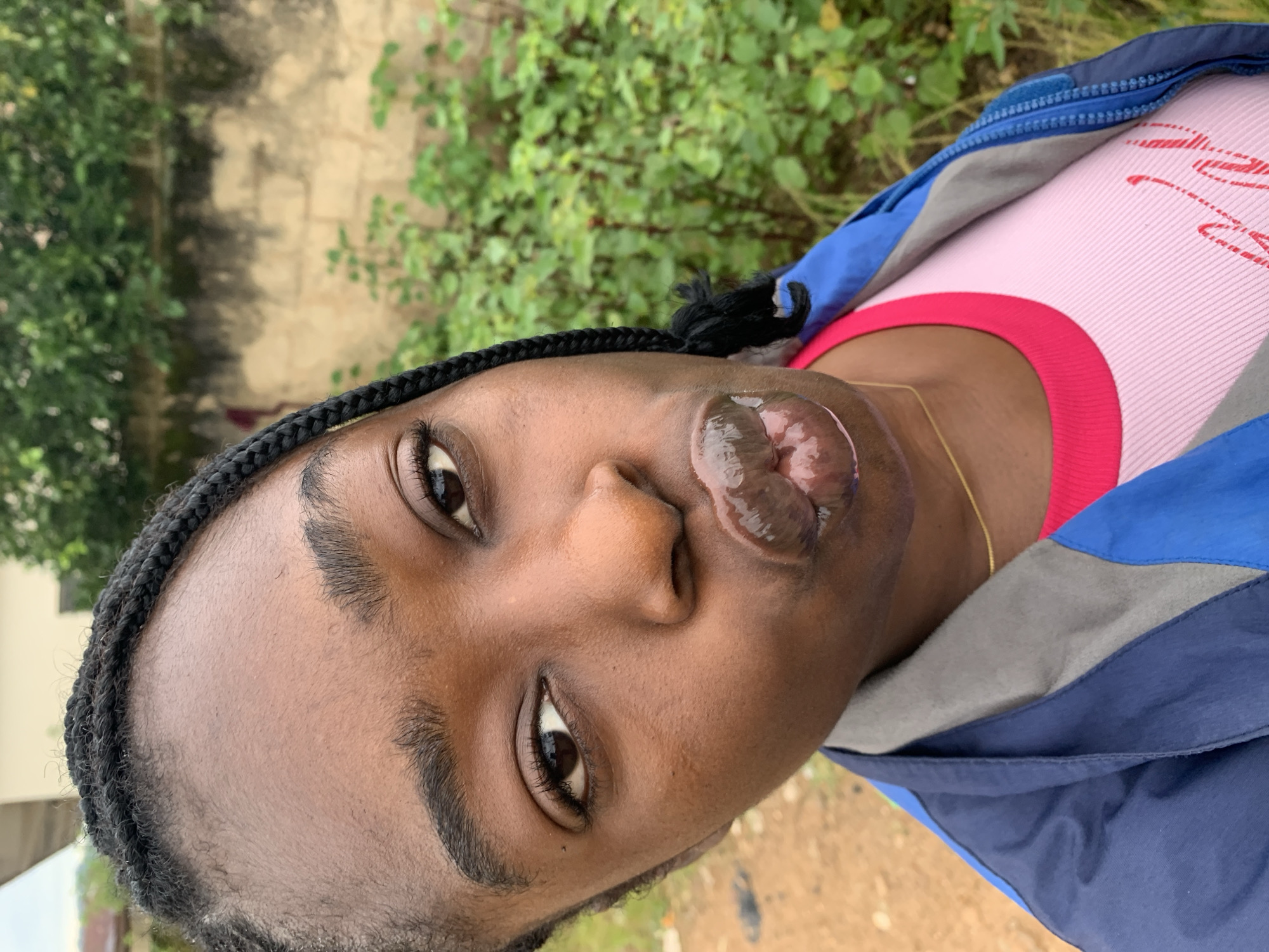 In an outdoor setting, likely in Rafin Par, Nigeria, a close-up shot focuses on the face of a young Black woman. She has dark skin, braided hair, and appears to be puckering her lips, which are shiny and appear to have lip gloss or balm on them. She is wearing a pink t-shirt with a red trim and a blue and grey jacket. The background shows greenery and a textured wall, suggesting a residential or natural environment.