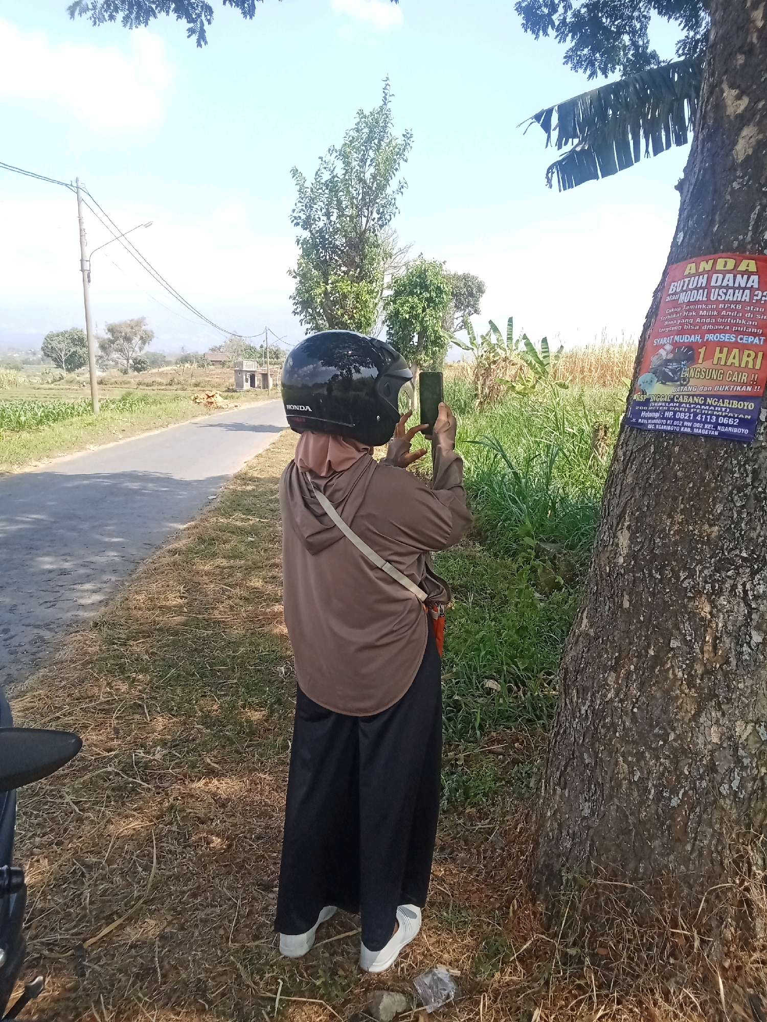 The image shows a person standing next to a tree in an outdoor setting, possibly in the city of Bulukerto, Indonesia, given the text on the poster. The person is wearing a brown hoodie, a headscarf, and dark pants, and is holding a smartphone to take a picture. A black helmet is on their head. The tree has a red and blue poster attached to its trunk with text written in Indonesian. The poster appears to be an advertisement for a loan or financial service, mentioning "ANDA BUTUH DANA" (Do you need funds?) and "MODAL USAHA" (Business capital). It also states "SYARAT MUDAH, PROSES CEPAT" (Easy terms, fast process) and "1 HARI LANGSUNG CAIR!!" (1 day, immediately disbursed!!). Below this, it lists the name "ASP TINGKAT CABANG NGARIBOYO" and an address and phone number. The background shows a paved road, a grassy field, and some vegetation, suggesting a rural or semi-rural area. The sky is clear and blue, indicating a sunny day.