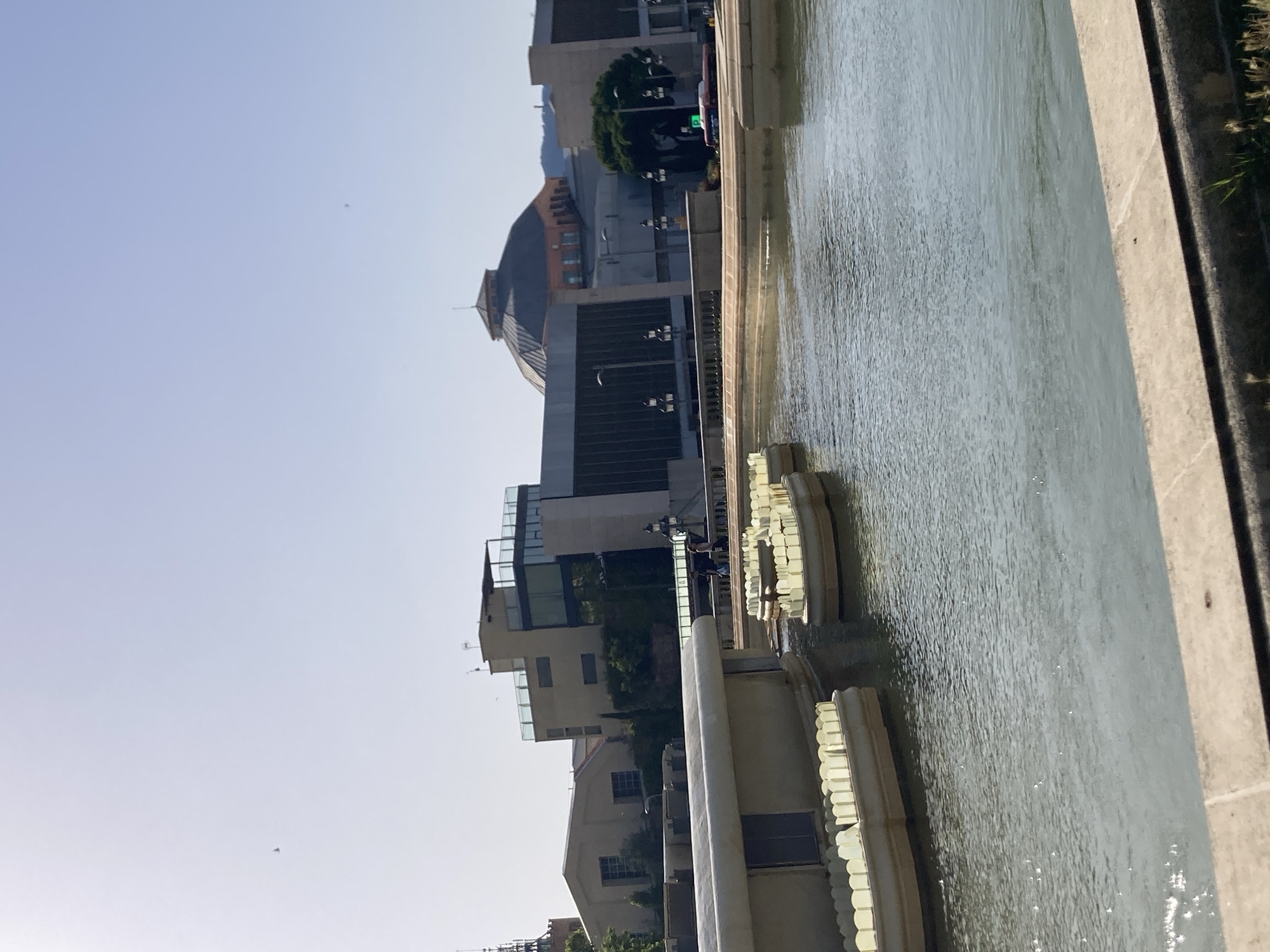 The image depicts a serene urban scene with a wide, calm body of water in the foreground. The water is bordered by a light-colored, stone embankment. Partially submerged in the water are several light-colored, boat-like structures, each with translucent, yellow vertical elements that appear to be decorative or functional lighting.
In the background, a collection of modern and contemporary buildings rises against a clear, pale blue sky. One prominent building features a large, domed roof with a segmented design and an orange-brick facade visible at its base. To the left of this dome, another building displays a more angular architectural style with glass facades and visible interior levels. Further to the left, a lighter-colored building with visible windows and a distinct roofline is present, and a slender, dark tower with a spired top is visible in the far left background.
Two individuals are visible walking on a raised walkway or bridge behind the water, in front of the buildings. The environment suggests a well-maintained public space, possibly a park or plaza, within a city. The sun is shining brightly, casting a gentle glare on the water's surface. The overall impression is one of architectural grandeur and open, public space on a clear day. The specific location is the Parc del Fòrum in Barcelona, Spain, which is known for its modern architecture and large water features.