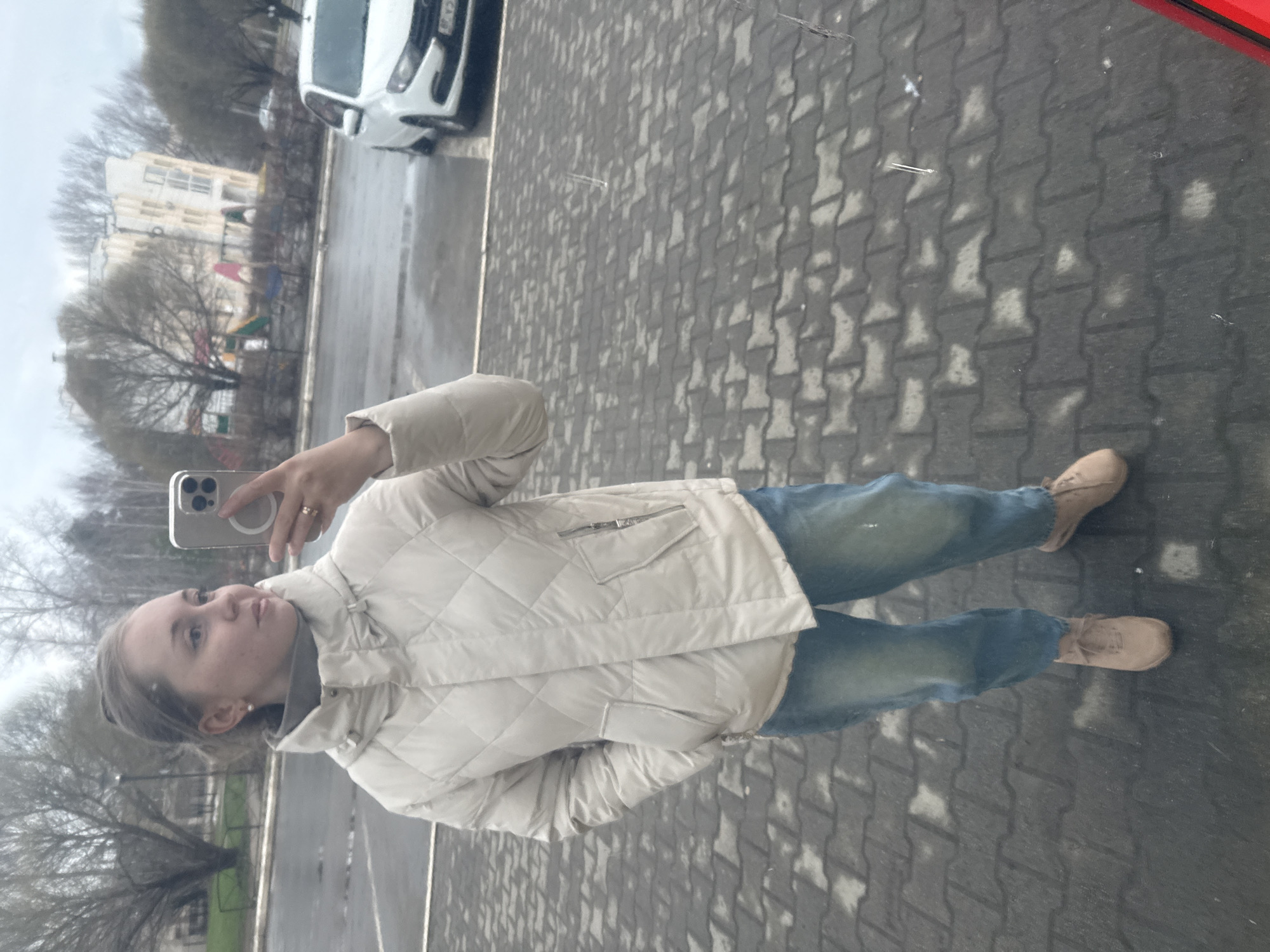The image is a reflection selfie of a young woman taken outdoors, likely in a large window or mirrored surface, indicated by the partial red frame visible in the top right corner and the overall reflective quality. The image is oriented sideways, requiring a 90-degree clockwise rotation for proper viewing.
The main subject is a young woman, seen from the waist up, holding a light-colored smartphone (possibly an iPhone) in her left hand to take the photo. She has light-colored hair pulled back and wears a light, possibly off-white or beige, quilted puffer jacket over a grey top, blue jeans, and tan lace-up shoes or boots. A ring is visible on her left ring finger, and she wears a small pearl earring in her right ear. Her expression is neutral as she looks towards the phone.
The reflected background shows an urban or suburban outdoor setting under an overcast sky, suggesting a daytime scene. Bare trees indicate late autumn, winter, or early spring. In the distance, a light-colored multi-story building and a children's playground with colorful equipment (red and blue structures) are visible. A road or street runs through the scene, with a white car (partially visible, possibly a Hyundai) reflected. The ground where the woman stands is paved with grey interlocking pavers.
The primary activity is the woman taking a selfie. The weather appears cool and overcast. A partial "M" is discernible on the reflected car's license plate; no other significant text is clearly visible.