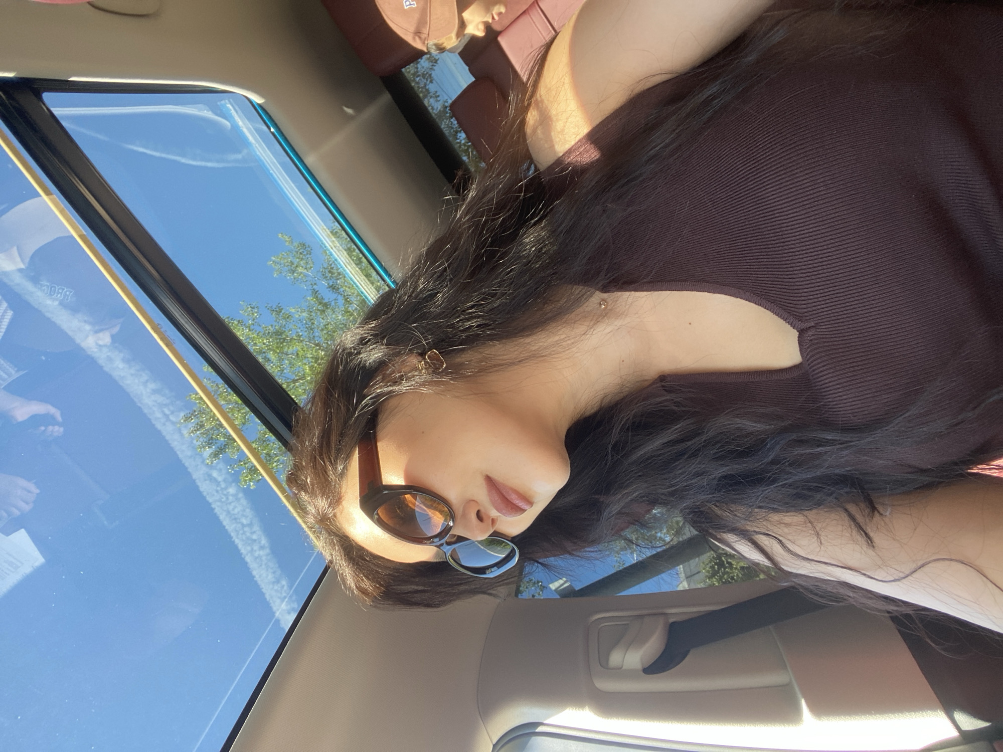 The image captures a person, likely female, with long dark hair and wearing sunglasses, inside a car in Sochi, Russia. She is depicted in profile, with her head slightly tilted, and is wearing a dark brown ribbed top.
The setting is the interior of a vehicle, prominently featuring a panoramic sunroof. Through the sunroof, a clear blue sky with distinct contrails is visible, indicating a sunny, daytime environment. Green tree foliage is also reflected in or seen through the glass, suggesting the car is outdoors. The bright sunlight casts highlights on her hair and skin.
Reflected in the sunroof, behind the main subject, a second person is partially visible, wearing a cap. The cap has text that appears reversed due to the reflection, with the letter "P" clearly seen, and other faint letters that could form a word. Additionally, the numbers "9087" are discernable in the reflections on the sunroof. The car's tan-colored headliner and a handle are also visible, completing the interior view. The scene conveys a relaxed atmosphere on a bright day.