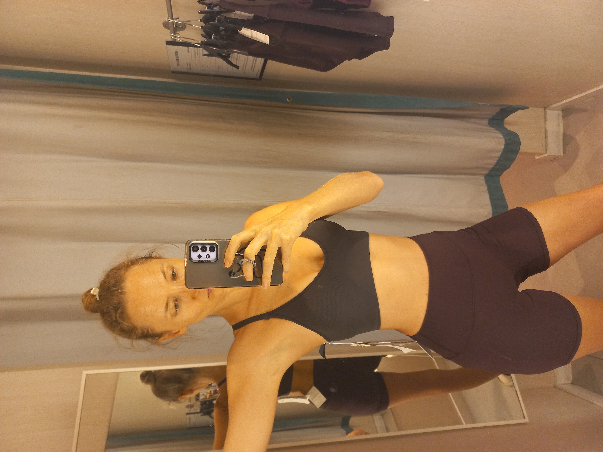 A woman takes a selfie in a dressing room in Pattaya City, Thailand, wearing a black sports bra and dark purple athletic shorts. She holds a phone in her right hand, capturing her reflection in a mirror in the dressing room. In the background is a curtain with a light blue wavy stripe near the top, and clothes hanging on a rack.