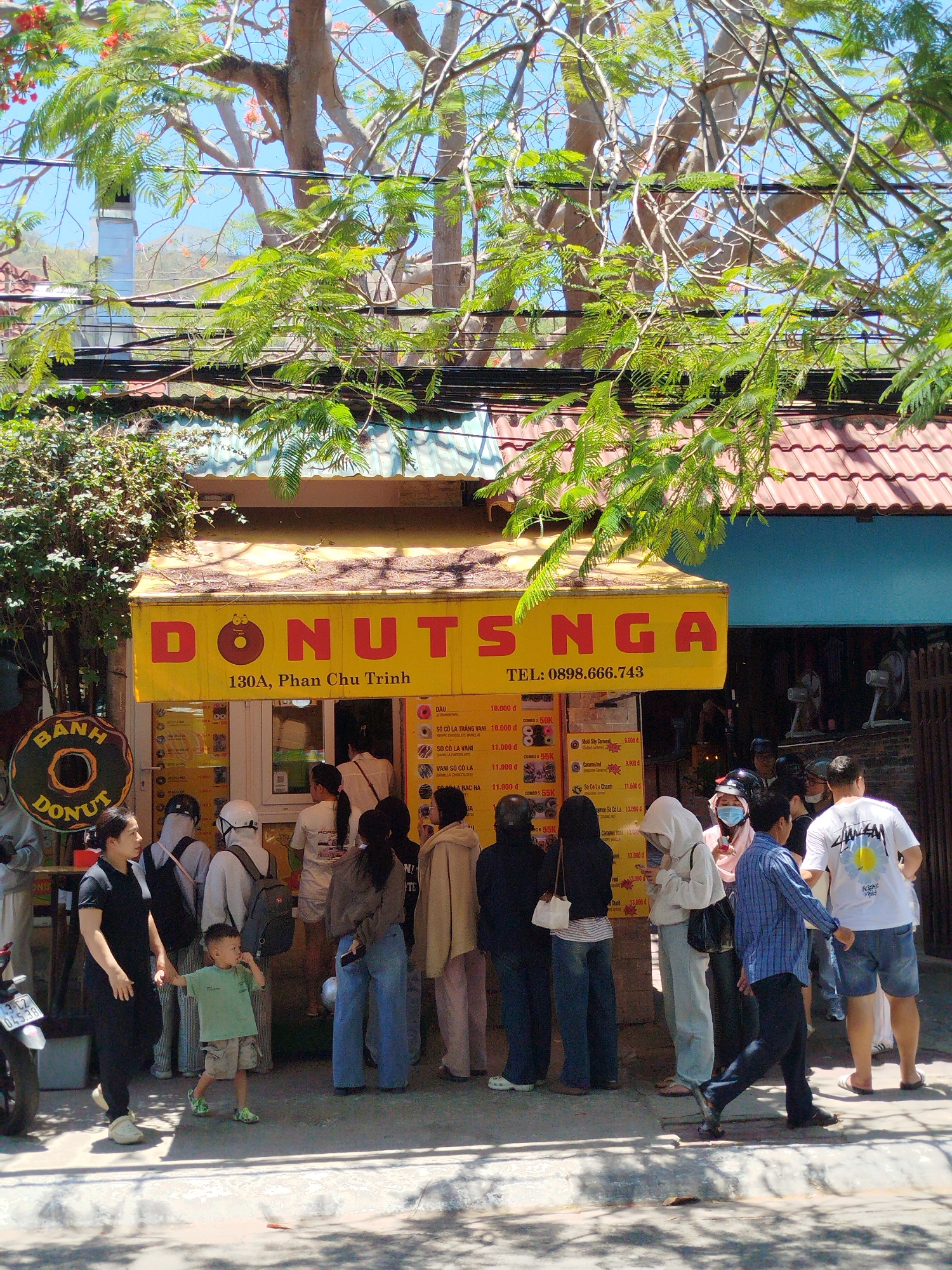 The image shows a bustling scene outside a donut shop named "Donuts Nga" located at 130A, Phan Chu Trinh in what appears to be Ho Chi Minh City, Vietnam, based on the street name. The shop has a yellow awning with the shop's name and phone number displayed.
A sizable crowd of people is gathered in front of the shop, presumably waiting to purchase donuts. They vary in age, dress, and gender, suggesting a diverse customer base. Some are wearing masks.
Signs displaying donut options and prices (in Vietnamese Dong) are visible, contributing to the local atmosphere. The surrounding environment includes trees providing shade, utility lines overhead, and building facades with tiled roofs, reflecting the urban landscape of Ho Chi Minh City. A parked motorcycle is visible to the left of the frame.