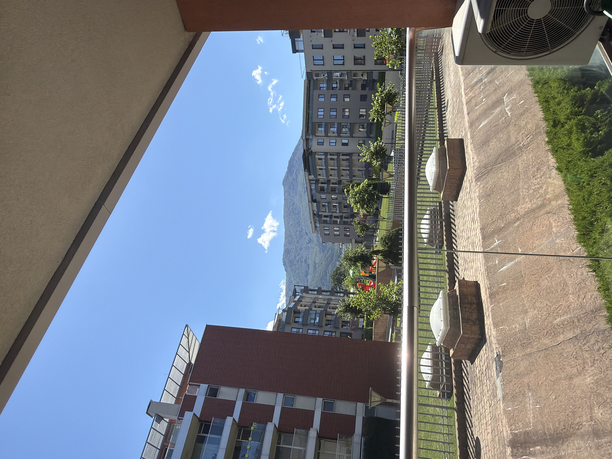 The image shows a view of a modern residential complex in Sofia, Bulgaria, on a clear day. In the foreground, there is a balcony with a metal railing and a large air conditioning unit. Beyond the balcony, a paved walkway leads to a green lawn with a playground. Several apartment buildings of varying heights and architectural styles are visible, with a backdrop of green, tree-covered mountains. The sky is bright blue with scattered white clouds.