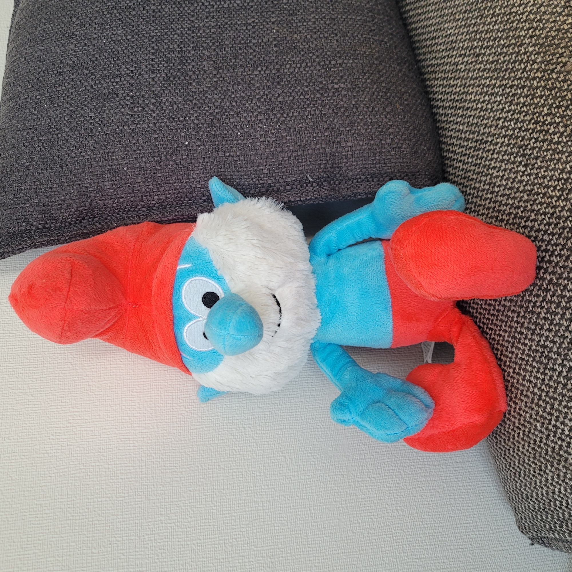A plush toy of a Smurf, dressed in red pants and a red pointed hat, sits on a textured gray sofa. The Smurf has blue skin, white fur around its face, and large, black eyes. The toy is positioned with its legs crossed and its arms resting on its lap. Behind the sofa, a textured white wall is visible. The overall scene is indoors, with a couch and wall typical of a residential setting. There is no indication of the location being in Jūrmala, Latvia.