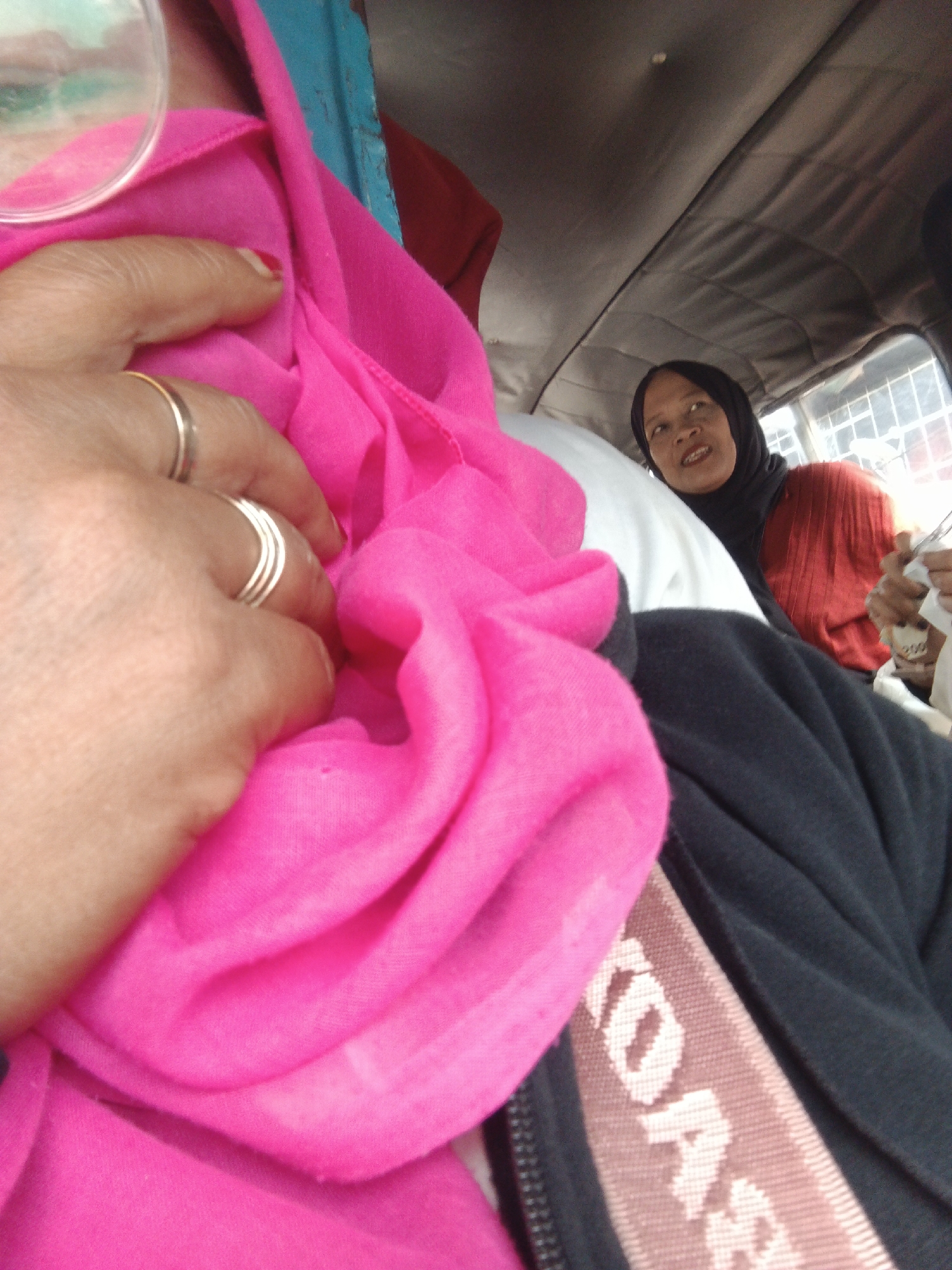 In Cikembar, Indonesia, a close-up shot reveals a woman's hand adorned with multiple silver rings, resting on a vibrant pink fabric. Beyond her hand, a woman in a black hijab and a red top is visible, looking upwards and to the side with an open-mouthed expression. The environment appears to be the interior of a vehicle, possibly an auto-rickshaw, with a textured grey ceiling and a bright blue element in the upper left corner. The overall lighting suggests it is daytime.