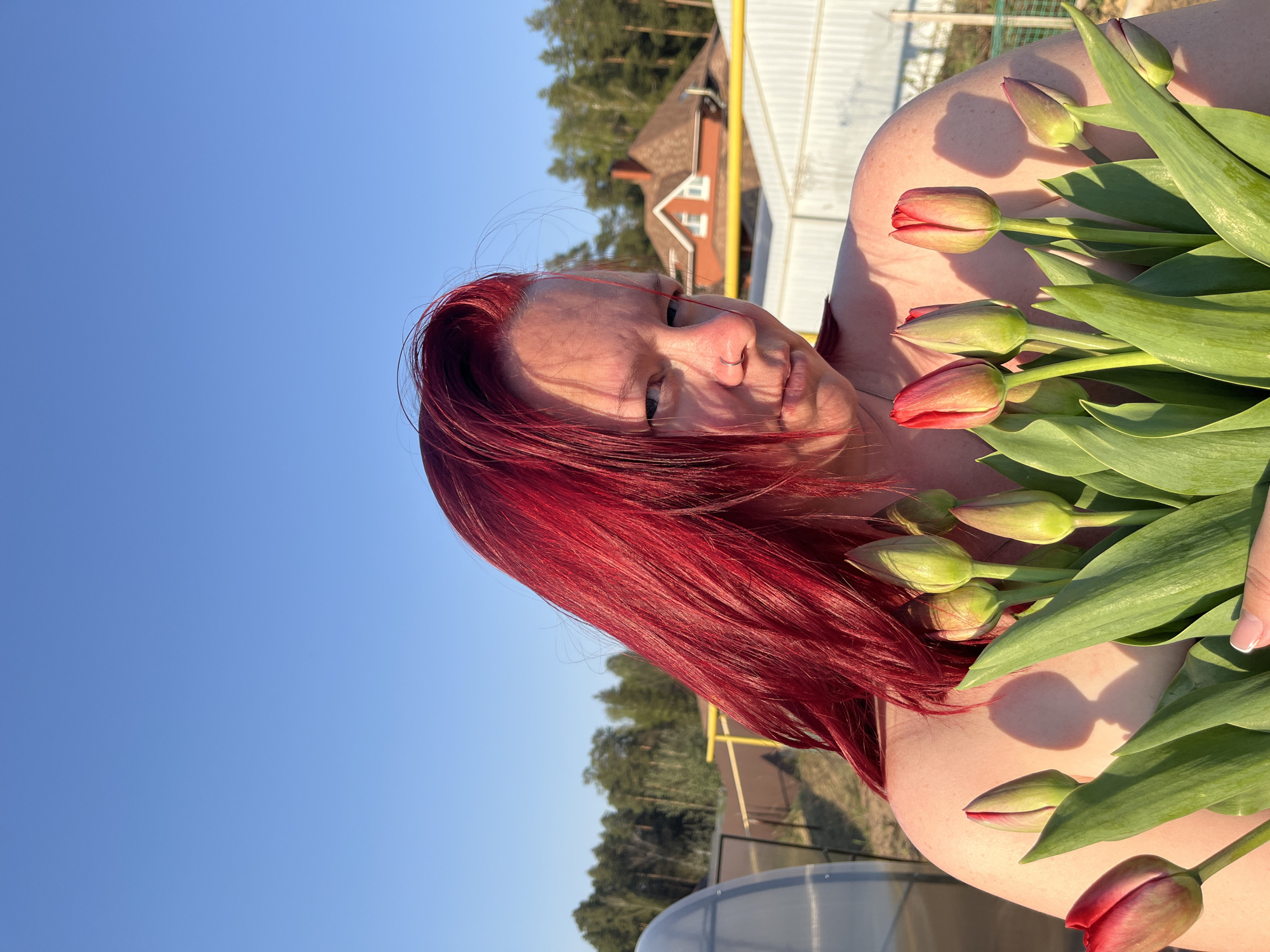 A woman with bright red hair holds a bouquet of tulips. She has a nose ring and is positioned at an angle in the shot, with the sky visible in the background. A building with a brown roof is partially visible behind her, along with trees. A metallic structure and the top of what appears to be a greenhouse are also visible in the lower portion of the frame. Based on the information provided, this image was taken in the city of Chaikovsky, Russia.