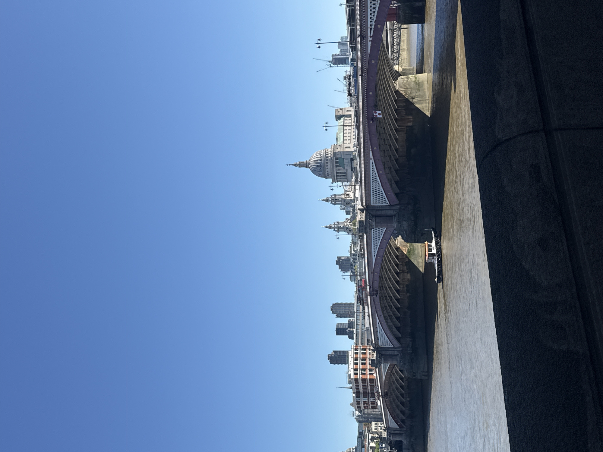 A photograph captures a wide-angle view of a city scene on a bright, clear day, with the perspective seemingly from a bridge or elevated structure. A vast, cloudless blue sky occupies approximately half of the frame, indicating excellent weather conditions. Below the sky, a large river flows, its murky brown water reflecting the light.
Spanning the river is a distinctive railway bridge with intricate red and white ironwork and multiple arches. Along the far bank, a dense urban landscape unfolds, featuring numerous buildings of various sizes and architectural designs. The most prominent landmark is the majestic dome of St. Paul's Cathedral, which stands out against the cityscape. Several cranes are visible in the distance, suggesting ongoing construction or development. A dark, textured wall or structure forms a substantial part of the foreground on the right side of the image, offering a stark visual contrast to the brighter elements. A small boat or barge can be seen navigating the river beneath one of the bridge's arches. The combination of St. Paul's Cathedral and a major river bridge clearly identifies the location as London, specifically looking along the River Thames, likely from a vantage point on or near Blackfriars Bridge. No discernible text is visible within the image.