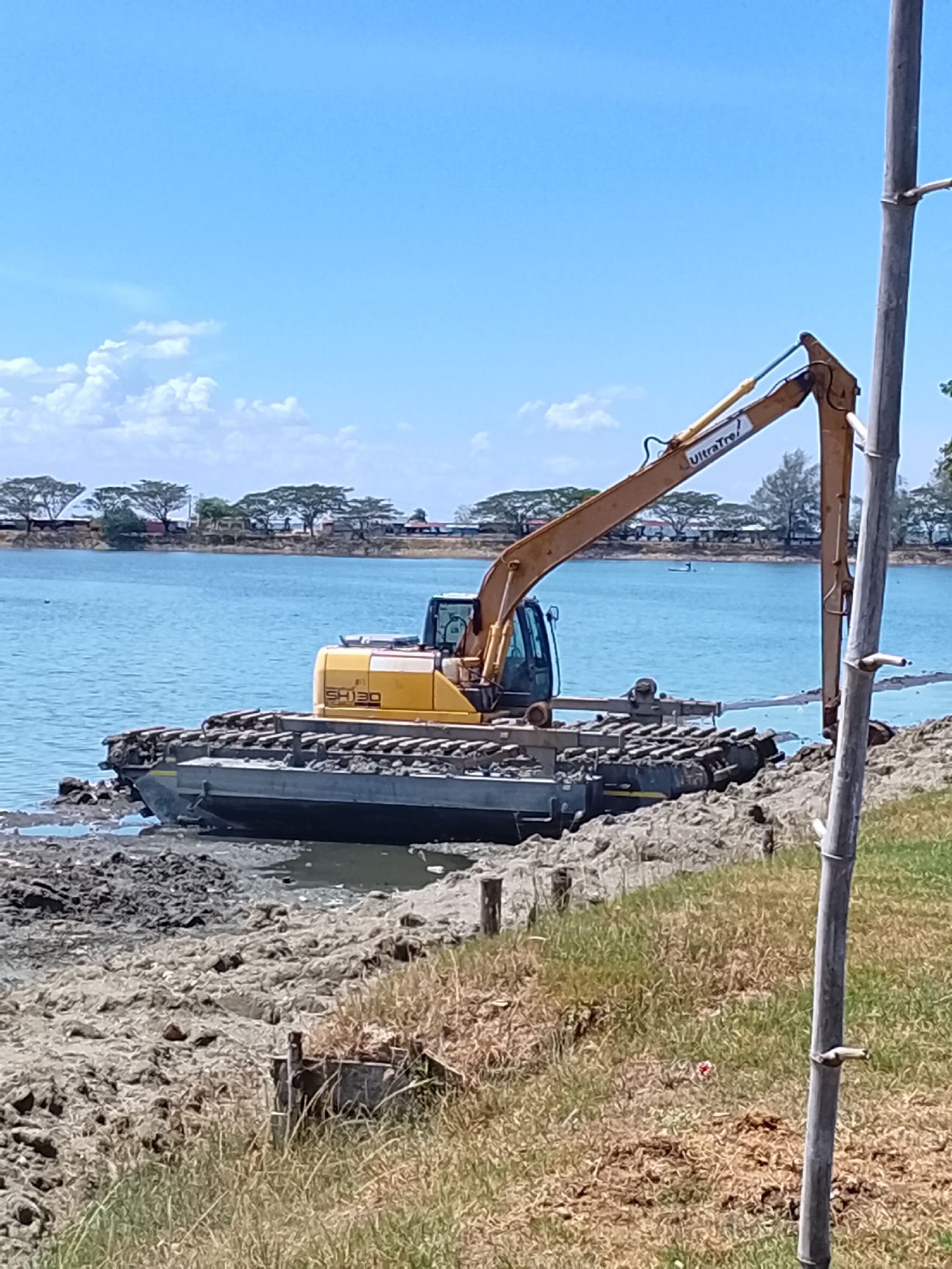 An amphibious excavator is partially submerged in the muddy bank of a body of water, with its tracks resting on both land and water. The excavator is yellow and gray, with "SH130" and "UltraTrec" visible on its arm. The water is a bright blue, and the sky is clear with scattered white clouds. In the background, trees line the far bank of the water, with buildings and some structures visible among them. The foreground consists of dry grass and mud. The scene is likely in Lhokseumawe, Indonesia, a city known for its coastal areas and industrial activities, where such heavy machinery might be used for dredging or construction.