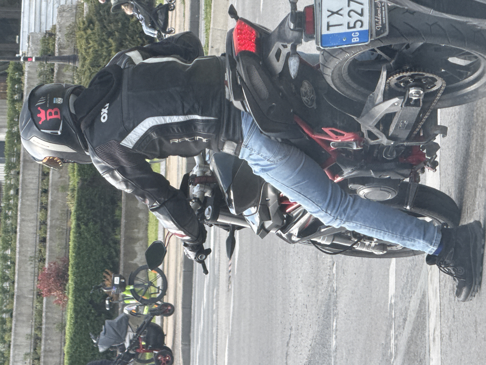 A person is depicted riding or stationary on a motorcycle on a paved road, captured in a dynamic, vertical shot. The rider is wearing full protective gear, including a dark helmet featuring a prominent red stylized "B" with a crown-like emblem, an "MT" logo, and the number "001". Their black leather motorcycle jacket has white and grey accents, with "OXI" visible on the shoulder and "RACING" printed on the sleeve. They are also wearing light blue jeans and black boots.
The motorcycle itself is dark, with visible red accents, particularly a bright red LED taillight. The rear wheel, chain, and sprocket assembly are clearly visible. A Bulgarian license plate is attached to the rear, displaying "TX BG 527". Below the main plate, a frame reads "Valdemi" with additional, unreadable small text.
In the background, a neat green hedge row separates the road from a concrete wall or fence. Further down the road, various objects resembling bicycles and a baby stroller can be seen. The scene is brightly lit, indicating it is daytime with clear weather. The setting appears to be a street or roadside in Varna, Bulgaria, as indicated by the "BG" on the license plate and the provided location context.
