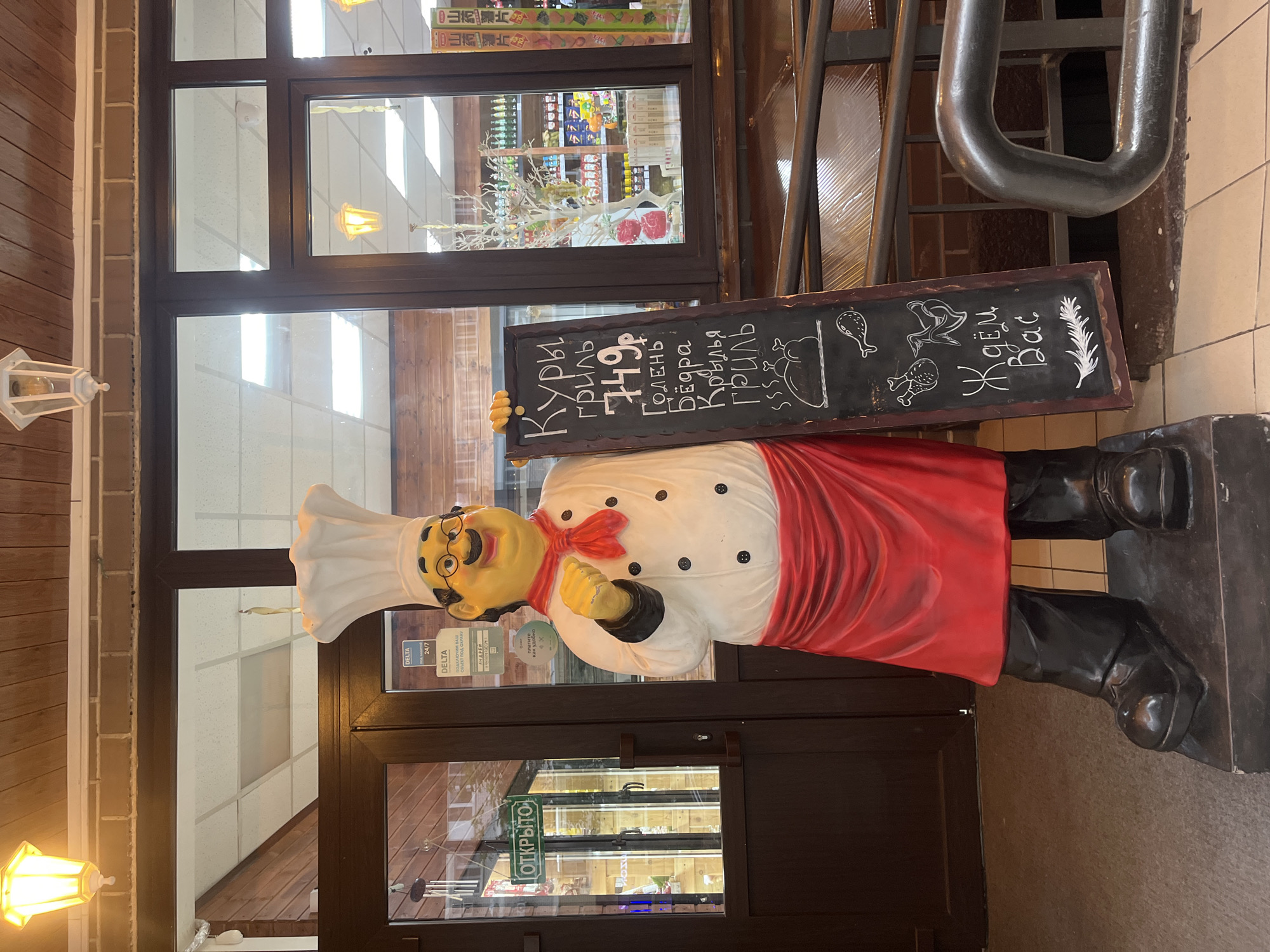 The image depicts a life-sized statue of a portly male chef, made of painted plastic or fiberglass, positioned at the entrance of a commercial establishment in Krasnodar, Russia. The chef wears a white toque, a white double-breasted chef's jacket with black buttons, a red neckerchief, a red apron, black pants, and black shoes. He has a yellow-skinned face with a red nose, round spectacles, and a black mustache. His right hand is raised to shoulder height, clenched, while his left hand holds a black chalkboard sign.
The chalkboard features white chalk writing in Cyrillic. The top text reads "Куры гриль" (Grilled Chicken), followed by "749р" (749 rubles). Below this, it lists "Голень" (Drumstick), "Бёдра" (Thighs), and "Крылья гриль" (Grilled Wings). Further down, there are chalk drawings of a whole roasted chicken, a chicken drumstick, and two chicken wings. The bottom text says "Ждём Вас" (We Await You). A feather drawing is at the very bottom.
The chef statue stands on a dark, elevated base. Behind the statue is a double-leaf glass door with dark brown frames, indicating an entrance to a building. To the left of the left door, a green rectangular sign with "ОТКРЫТО" (OPEN) in white Cyrillic is visible. Various security stickers from "DELTA" (likely a security company) are affixed to the glass of the right door, including one that mentions "24/7" and another stating "ПОД ОХРАНОЙ" (UNDER GUARD). Another sticker features text "АНДЕЕ" and "платите как удобно" (pay as you like).
Through the glass doors and windows, an interior space with bright overhead fluorescent lighting and a suspended ceiling with square panels is visible. The interior also shows shelves stocked with various products, suggesting a convenience store or grocery section. A decorative white tree with red berries is visible inside, near the window. The exterior overhead features a wooden slatted ceiling, brickwork, and several wall-mounted lanterns emitting a warm yellow light. The ground level visible around the statue is a dark, possibly carpeted or tiled, surface. A dark metal railing is partially visible on the right, suggesting a ramp or steps leading to the entrance. The overall scene suggests a commercial establishment, likely a restaurant or a shop selling ready-to-eat grilled chicken.