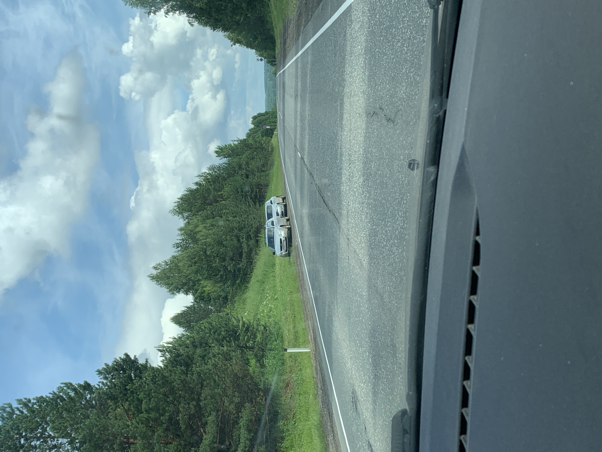 The image shows a view from inside a car looking out at a road in the city of Sudak, Russia. The road is asphalt and has white lane markings. On the right side of the road, there is a grassy embankment with trees. On the left side of the road, there are two silver cars parked on the shoulder. One car is a hatchback, and the other appears to be a station wagon. The sky is blue with white clouds, and the sun is shining. There are trees and hills in the distance. The overall scene suggests a scenic drive on a summer day.