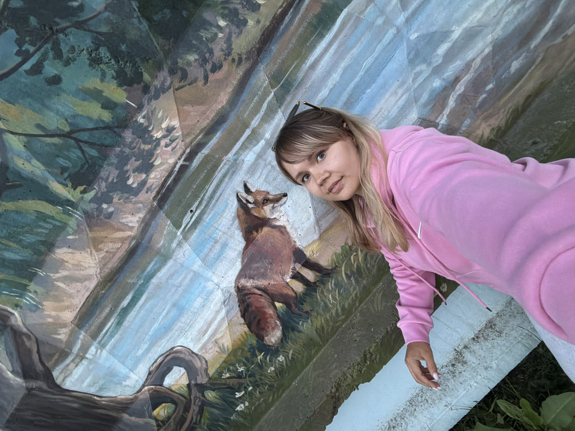 A young woman with blonde hair and bangs, wearing a pink hoodie, poses for a selfie in front of a large mural. The mural depicts a natural scene with a river, trees, and a fox. The woman is positioned on the right side of the frame, with her left arm extended to take the photo. She is seated on a white bench, with green foliage visible at the bottom left of the image. The mural appears to be painted on a curved surface, possibly a section of a stadium or outdoor seating area. The mural is likely located in the city of Chaikovsky, Russia, as indicated by the context.