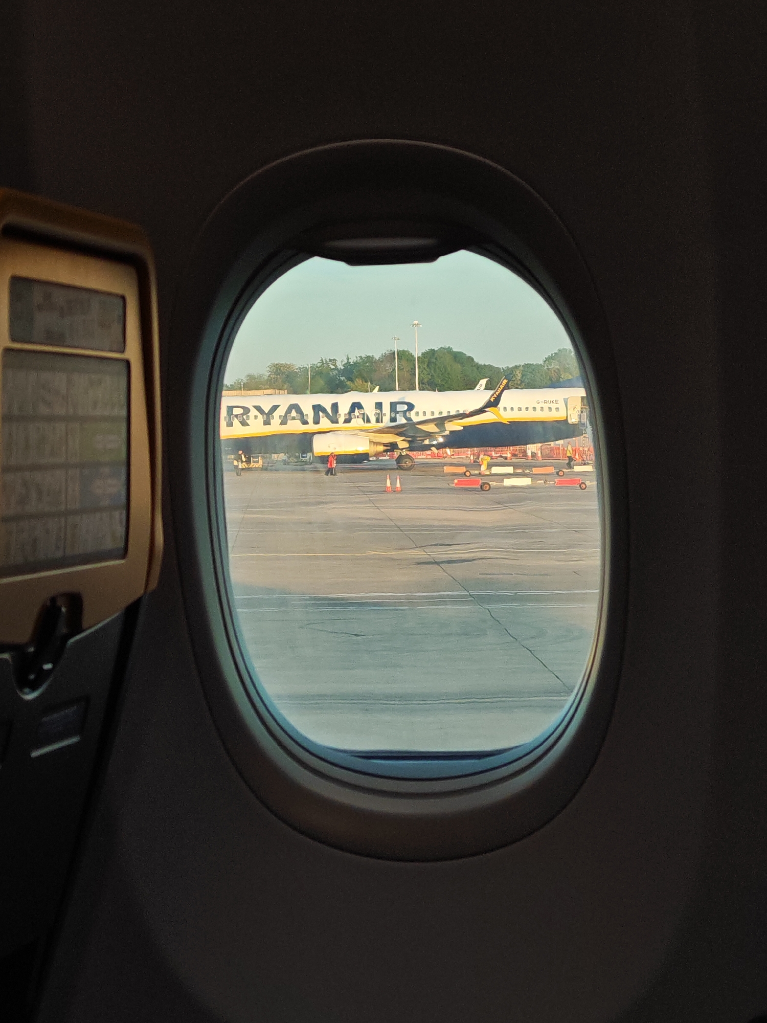 From the perspective of an airplane passenger looking out the window, a Ryanair aircraft is visible on the tarmac. Ground crew are working around the plane. The registration G-RUKE is visible on the aircraft. The location is likely at an airport within Uttlesford, United Kingdom. The interior of the passenger plane with a foldable tray and interior panel is also visible.