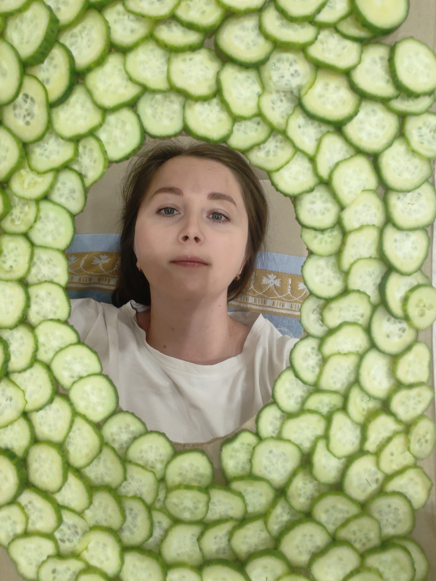 A medium-shot photograph shows a young woman in a white t-shirt lying down, framed by an oval border of sliced cucumbers. She has fair skin, light brown hair, and is looking directly at the camera with a neutral expression. Her head rests on a light blue and beige patterned surface, possibly a pillow or cloth. The cucumber slices are arranged densely around her face and upper body, creating a frame. The location is Unknown, Unknown.