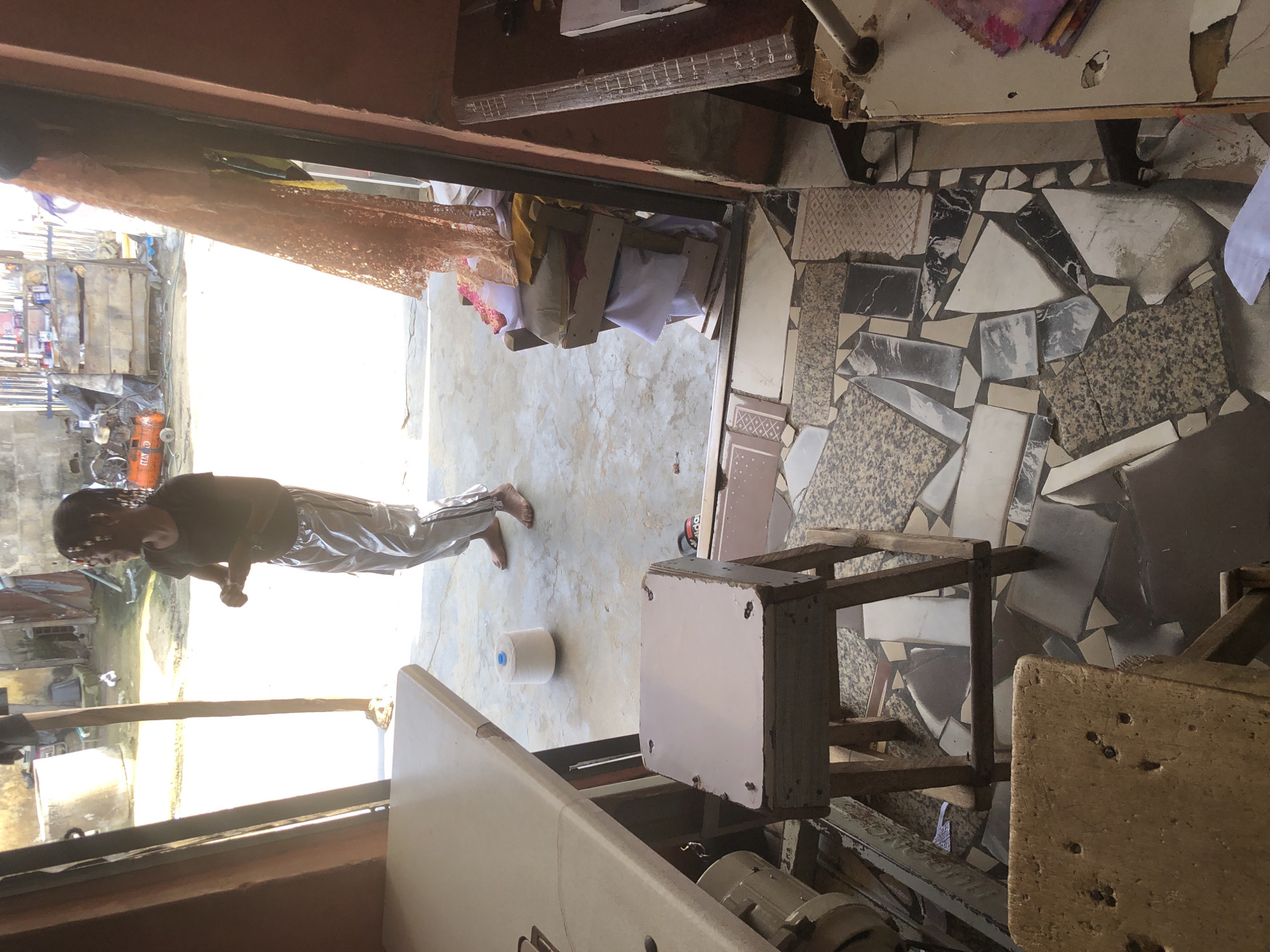 In Ado, Nigeria, a young Black woman with her hair in beaded braids stands in the doorway of a sewing workshop. She wears a dark t-shirt and shiny silver pants. Her hands are clasped in front of her. She is looking to her right, out of the workshop and into a bright, open area. The workshop is cluttered with sewing equipment, stacks of fabric, and a wooden stool. The floor is covered in a mosaic of broken tiles, some of which are a dark gray marble pattern, others a lighter beige with a diamond pattern, and some are solid white or beige. In the background, a concrete structure and some metal shelves with various items are visible. To the right of the woman, inside the workshop, a roll of white thread sits on the floor. A sewing machine and table are in the foreground on the left.