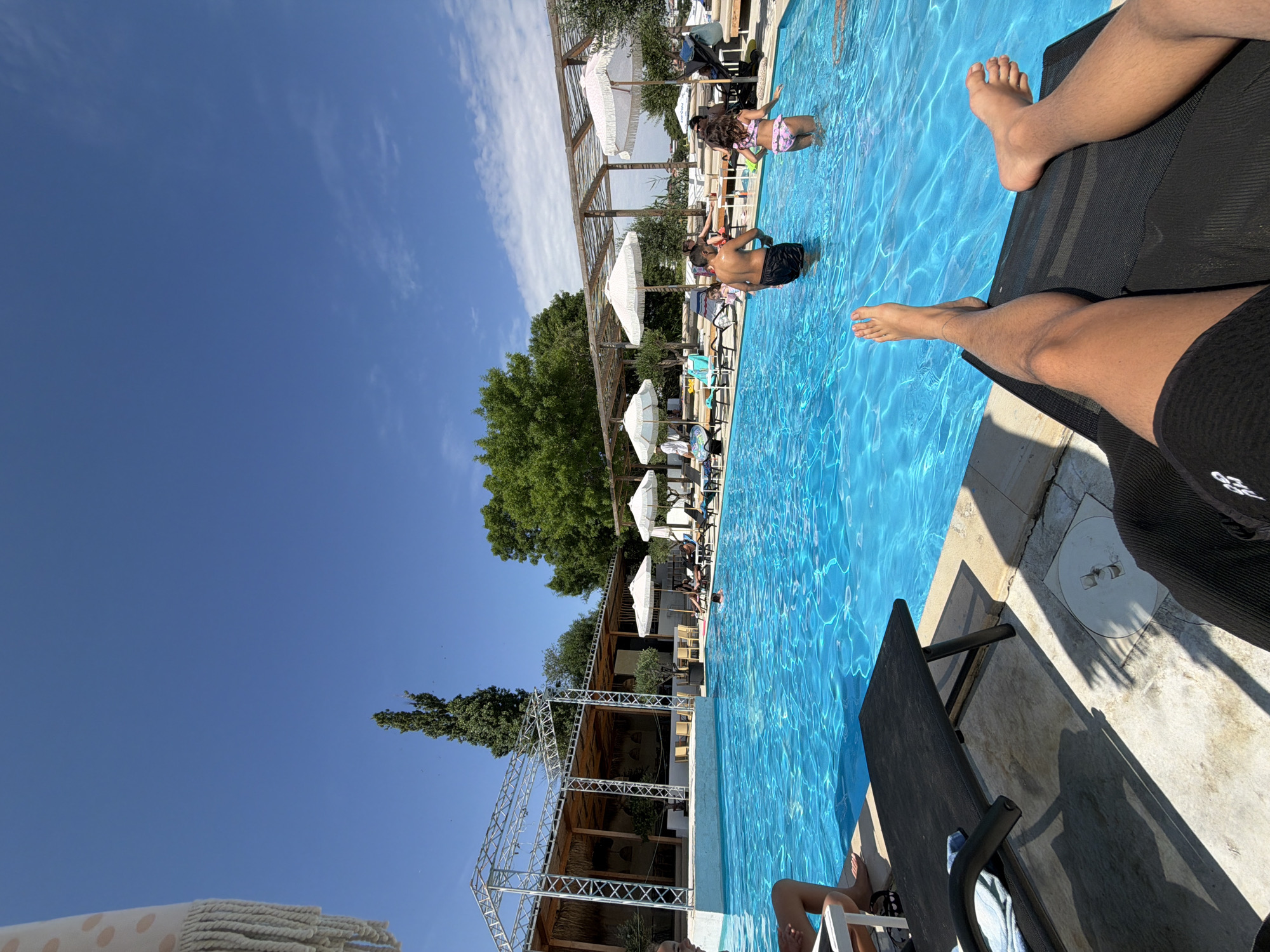 The image depicts a poolside scene on a sunny day, likely at a resort or hotel in Burgas, Bulgaria. In the foreground, a person's legs are visible, stretched out on a black lounge chair. The pool itself is filled with vibrant blue water, with several people visible. A man and a young girl are standing in the water, seemingly interacting. Other individuals are scattered around the pool area, some lounging on chairs or in the water. Behind the pool, there are lounge chairs with white umbrellas, a thatched-roof structure, and lush green trees. The sky is a clear, bright blue with a few wispy clouds. The overall atmosphere is relaxed and recreational.