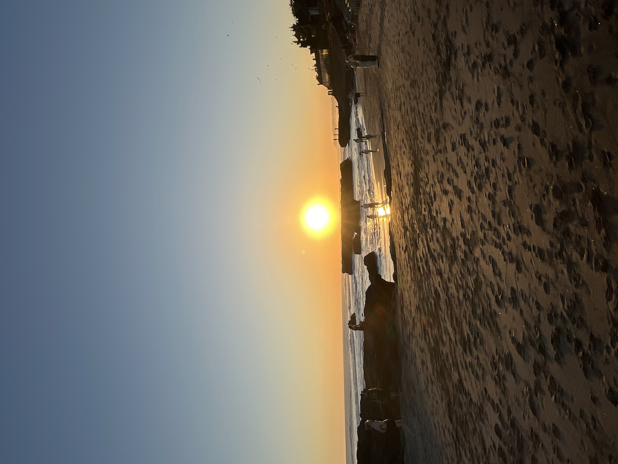 Captured during the golden hour, the photograph shows a seaside scene in North Kuta, Indonesia, dominated by warm colors from the setting sun. The sky transitions from a light blue to a bright golden-yellow where the sun dips close to the horizon. Silhouetted figures are walking along the shoreline. The beach is dark brown and textured with footprints. The ocean reflects the sunlight. Near the foreground, a silhouette of a person appears to be standing on a rock formation.