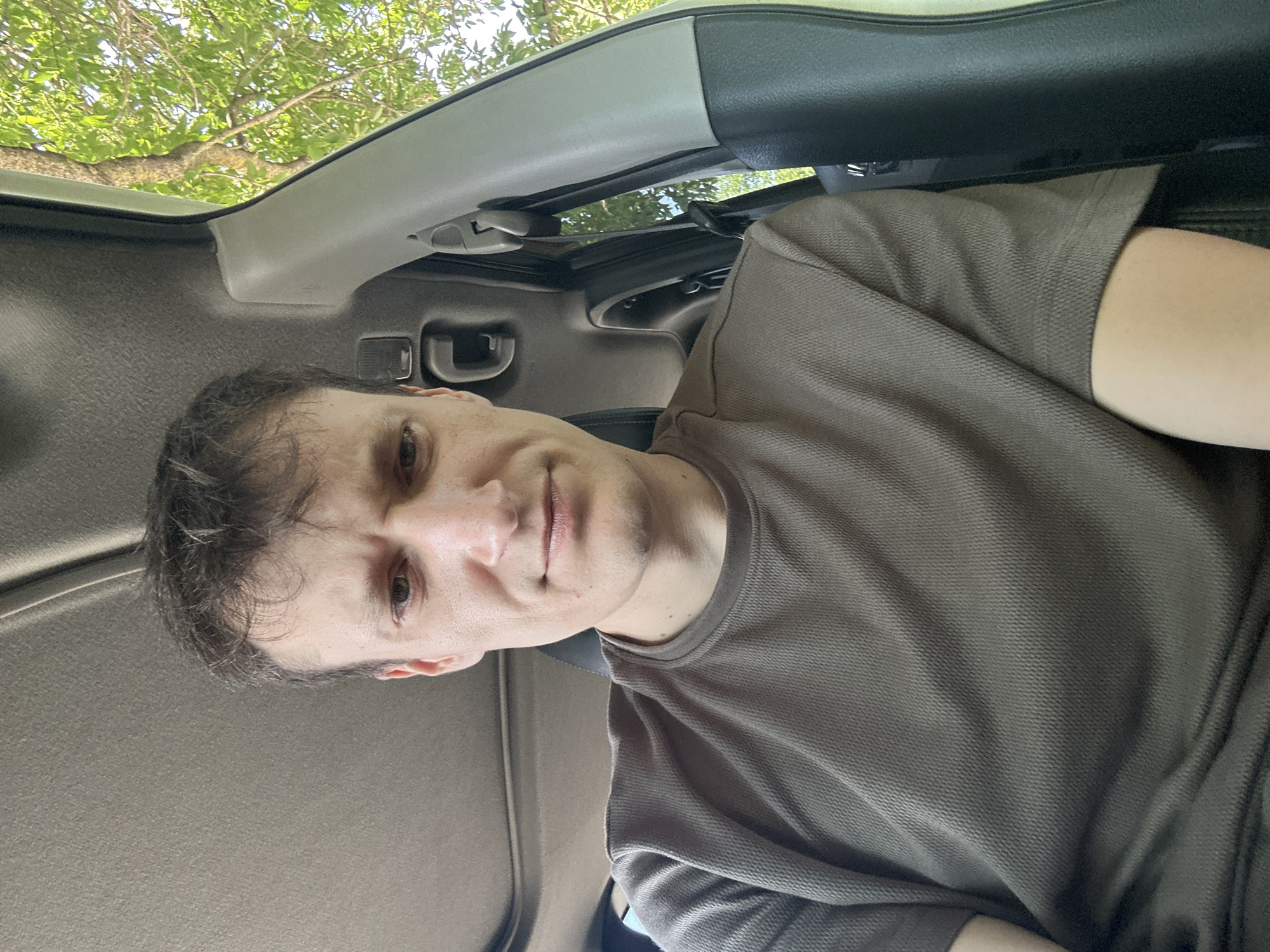 A man is sitting in the driver's seat of a car, looking at the camera. He has short, light brown hair and is wearing a brown t-shirt. The interior of the car is visible, including the headliner and a part of the dashboard. Outside the car, green trees and sunlight are visible through the window. The image appears to have been taken on a sunny day. The location is not specified to be in the city of Makiv, Ukraine.