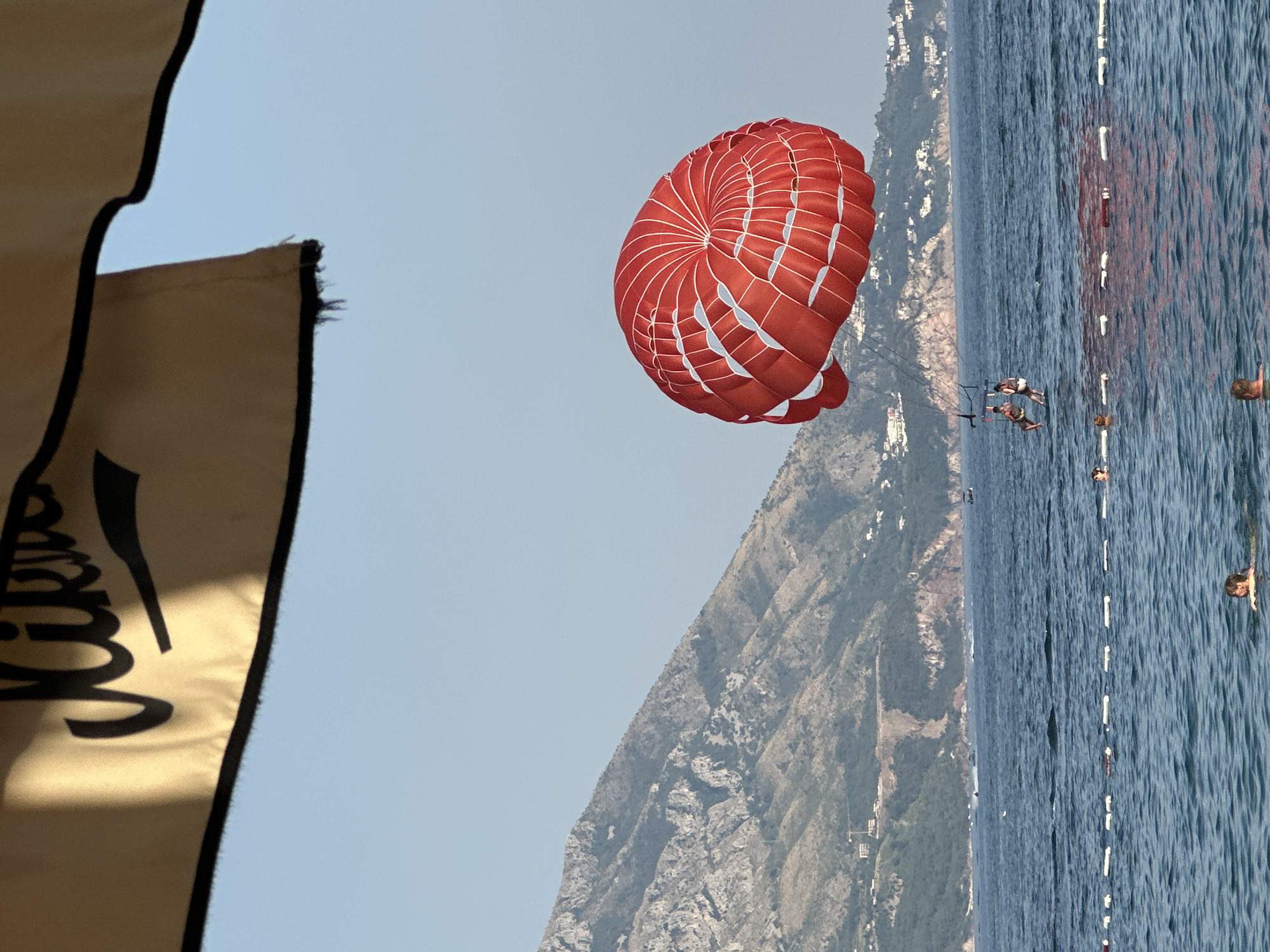 The image shows a parasailing activity on a sunny day in Budva, Montenegro. A bright red and white parachute is prominently featured in the sky, with several people visible in the water below, some appearing to be parasailing or enjoying water activities. The background consists of a steep, rocky mountain covered in lush green vegetation, with scattered buildings suggesting a coastal town. The sea is a deep blue, with a line of buoys marking an area in the water. The overall scene evokes a sense of leisure and adventure. A dark flag with black lettering is partially visible in the foreground, suggesting it might be associated with the parasailing operation. The sky is clear and blue, indicating good weather conditions. The time of day appears to be daytime, likely late morning or afternoon, given the bright sunlight.