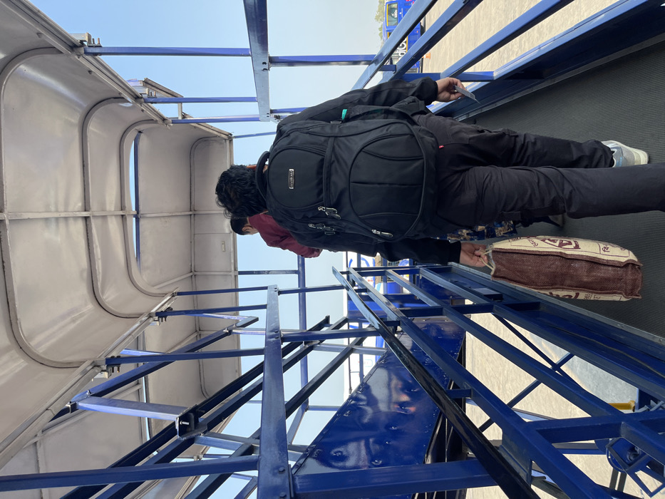 The image, taken in Guwahati, India, captures a moment of people moving along a metal ramp or stairway, likely at an airport. The scene is viewed from a low angle, looking upwards towards a clear blue sky.
In the foreground, a person is seen from behind, walking away from the viewer. This individual wears a black jacket, black pants, and light-colored athletic shoes. A black backpack with "SKYBAGS" visible on a small tag is worn on their back. In their right hand, they hold a small white card or ticket. Their left hand holds a brown and white patterned tote bag, on which the letters "bal" and "Kg" are discernible, suggesting it's a grocery or utility bag.
Behind this main figure, a second person is partially visible, also seen from behind, wearing a maroon top. A third person is faintly seen further back. The people appear to be ascending or descending a dark, flat walkway supported by a prominent blue metal framework of stairs and beams. To the left, a light-colored, curved metallic structure, resembling a gangway or covered walkway, frames the upper part of the scene. In the distance, beyond the structures, faint outlines of what appear to be airport ground vehicles or other aircraft are visible under the bright, clear sky.
The weather is bright and sunny, indicating daytime. The activity suggests passengers in transit, possibly boarding or disembarking.