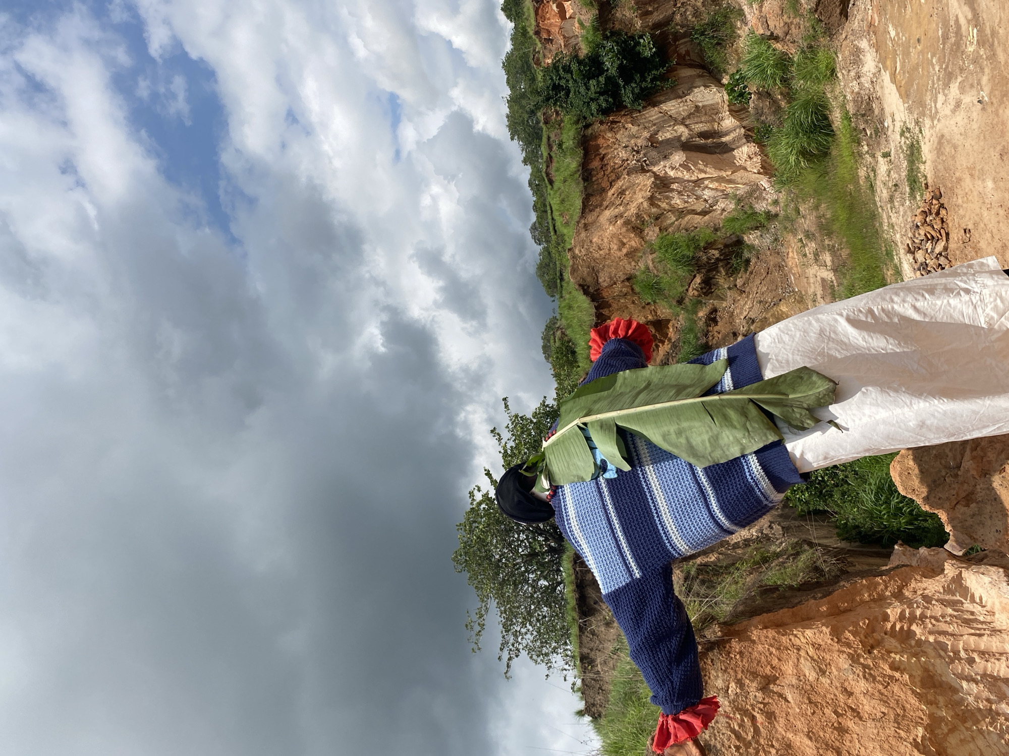 The image shows a person standing in a rural, outdoor environment, likely a quarry or a steep, eroded hillside, in Ufu Esioni, Nigeria. The person is seen from behind, facing away from the camera and towards a dramatic sky filled with both dark, heavy clouds and patches of blue sky. The individual is wearing a blue and white striped sweater, a black head covering, and a long, flowing off-white garment. Red fabric is visible on the cuffs of the sweater, and a large green banana leaf is attached to the back of the sweater. The landscape is characterized by reddish-brown soil, exposed rock faces, and sparse green vegetation. The terrain appears uneven and rugged, suggesting a natural or semi-developed area.