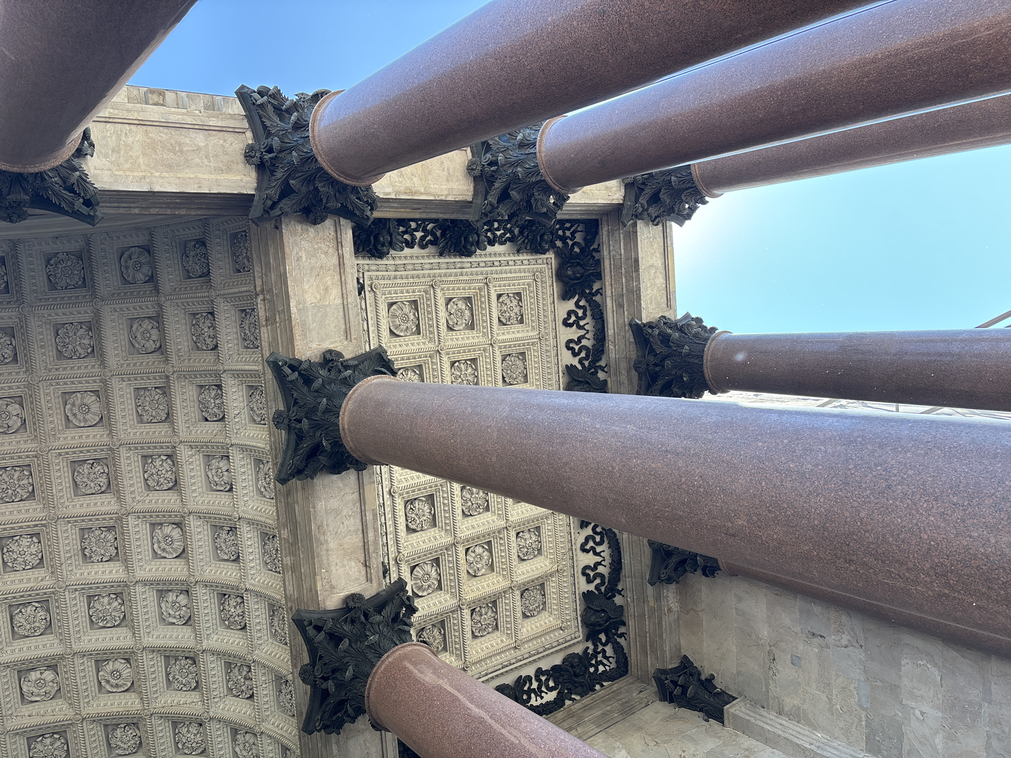 This is a low-angle, upward shot of the colonnade of Saint Isaac's Cathedral in Saint Petersburg, Russia. The image features massive, polished red granite columns supporting a richly ornamented white ceiling. The ceiling is a grid of square coffers, each containing a decorative floral relief and surrounded by intricate molding. Dark, ornate capitals top the columns, and further decorative elements are visible along the underside of the portico. The sky, a clear, bright blue, is visible through the gaps between the columns. The scene is devoid of people.