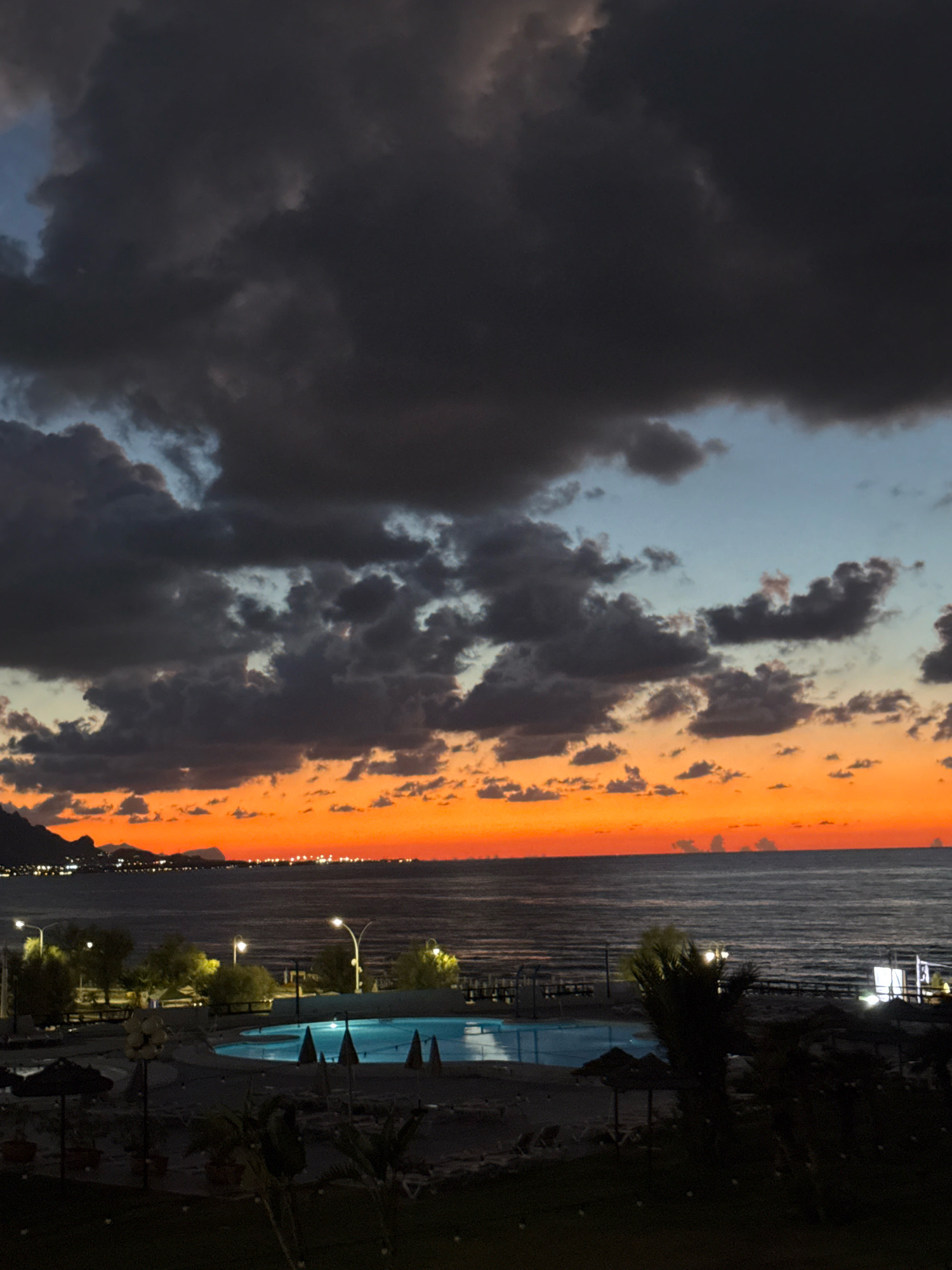 The image depicts a coastal scene at dusk, likely in Isola delle Femmine, Italy, given the context. The sky is dominated by dramatic, dark clouds that contrast with a vibrant band of orange and red hues at the horizon, indicating the setting sun. Below the sky, the sea is visible, appearing calm with subtle ripples.
In the foreground, a resort or hotel area is dimly illuminated. A bright blue swimming pool is a prominent feature, with its water reflecting the twilight sky. Surrounding the pool are loungers, umbrellas, and scattered vegetation, including palm trees. Several lampposts are also visible, casting pools of light. In the distance, along the coastline, a town or city is lit up with numerous lights, suggesting a populated area. The silhouette of hills or mountains can be seen on the far left, further defining the landscape. No people are clearly discernible in the image. The overall atmosphere is one of quietude and natural beauty as day transitions to night.