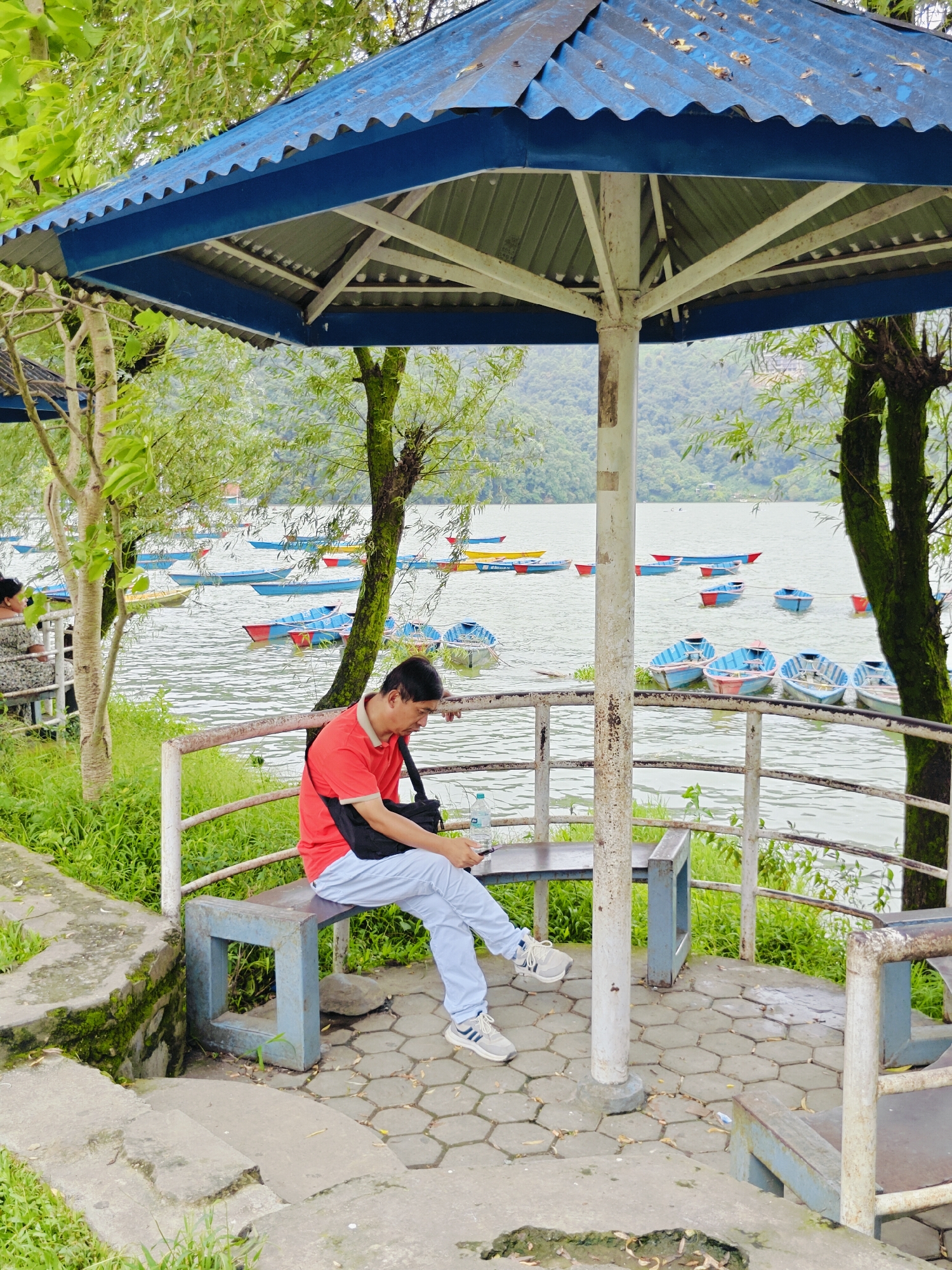 A man in a red polo shirt and light blue pants sits on a bench under a blue, corrugated metal awning. He is looking down at a cell phone or tablet in his hands. The bench is part of a covered outdoor seating area with a view of a lake. Several colorful boats are docked on the lake, and a lush green hillside is visible in the background. The scene appears to be in Pokhara, Nepal, likely near Phewa Lake, given the presence of boats and the surrounding natural landscape.