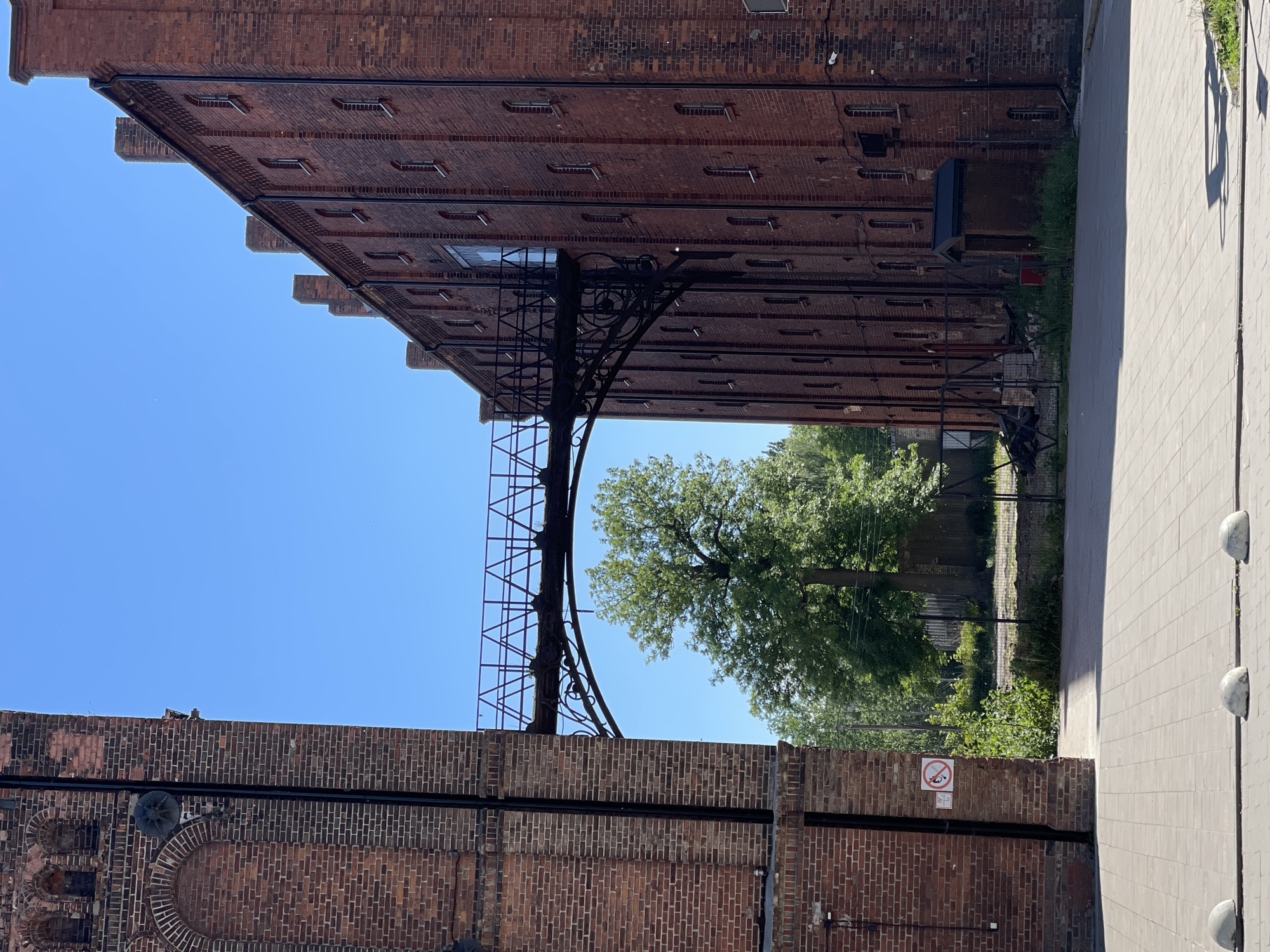 This image presents a detailed view of an industrial or historical urban setting in Kaliningrad, Russia, characterized by two tall, red-brick buildings connected by an elevated metal walkway.
The building on the left, partially visible, is constructed from dark red brick with darker mortar lines. It features several arched recesses in its upper section, consistent with former window openings or decorative elements. A circular, spoked metal object is mounted high on its facade. A black vertical pipe runs down the building's side. Near the bottom left, a white circular sign with a red border and a diagonal line crossing through a figure on a board indicates a prohibition against skateboarding or similar wheeled activities. Below it is a smaller, illegible white sign. The ground in the immediate foreground is paved with light gray square concrete pavers, with three white, hemispherical concrete bollards positioned along a joint line.
The building on the right is a much taller, longer structure, also made of dark red brick. Its facade is punctuated by numerous narrow, arched windows, arranged vertically in columns. The roofline features several prominent, rectangular brick chimney-like extensions. Black vertical pipes run down this building's exterior as well. At ground level, near the base of the right building, a small, dark brown, shed-like structure with a black roof is visible, surrounded by overgrown green foliage and situated behind a chain-link fence. A black security camera is mounted on the building's upper wall.
The elevated walkway connecting the two buildings is made of dark metal. It features a simple open-truss railing structure and is supported by an ornate, curved metal framework on its left side, connecting to the shorter building.
Beyond the buildings and the walkway, a vibrant natural background is visible, dominated by several large, mature trees with dense green foliage. A single prominent tree stands centrally beneath the elevated walkway. The sky above is clear and bright blue, indicating fair weather. Strong sunlight casts distinct shadows on the paved ground in the foreground. No people are present in the scene.