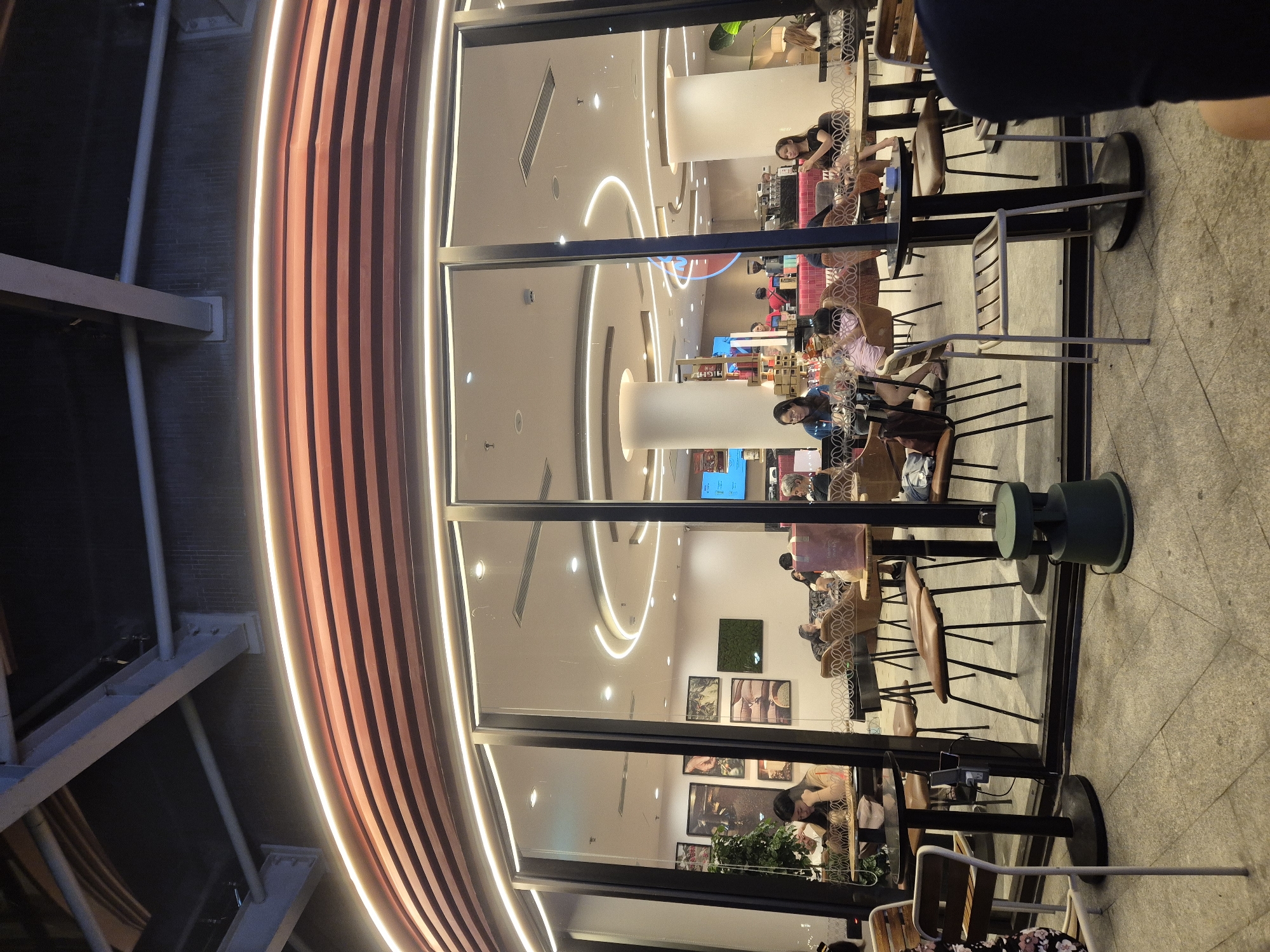 The image shows the interior of a modern cafe, viewed from the outside. The cafe has a bright and contemporary design, with sleek furniture and decorative lighting. The lighting fixtures inside are a prominent feature, with curved neon-like lights illuminating the ceiling and pillars. Several people are visible, seated at tables and chairs, some appearing to be customers and others possibly staff. They are engaged in various activities such as eating, drinking, talking, and working on their devices.
The cafe is situated in a location that suggests it is either in a mall or a busy urban area, given the external architecture and the presence of other structures visible in the background. The time of day appears to be evening or night, as the interior lights are brightly lit against a darker exterior, and the reflections on the glass suggest ambient light from outside is minimal. The overall atmosphere seems relaxed and inviting, typical of a cafe setting where people gather to socialize or work.
There are several framed pictures or artworks displayed on the walls inside the cafe, adding to the decor. Some of the tables have menus or other items on them. The seating arrangements include a mix of individual chairs and larger table setups. The flooring inside the cafe appears to be a light-colored tile. Outside the cafe, there is a paved area with more tables and chairs, suggesting an outdoor seating option. The exterior of the building features a curved facade with vertical wooden slats and integrated lighting, creating a striking visual element. The image is oriented vertically.