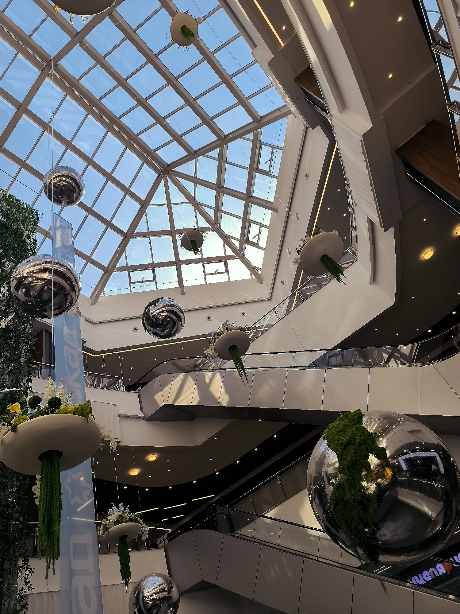 The image shows the interior of a modern shopping mall, likely in the city administration of Qarağandı, Kazakhstan, given the context. The multi-level atrium features a large glass ceiling, allowing natural light to illuminate the space. Suspended from the ceiling are several large, spherical metallic decorations and unique hanging planters filled with greenery and flowers, resembling giant turnips with cascading moss. Escalators are visible on different levels, indicating the flow of shoppers. The architecture is contemporary, with clean lines, white surfaces, and some wooden accents. The overall impression is one of a bright, spacious, and aesthetically decorated commercial environment.