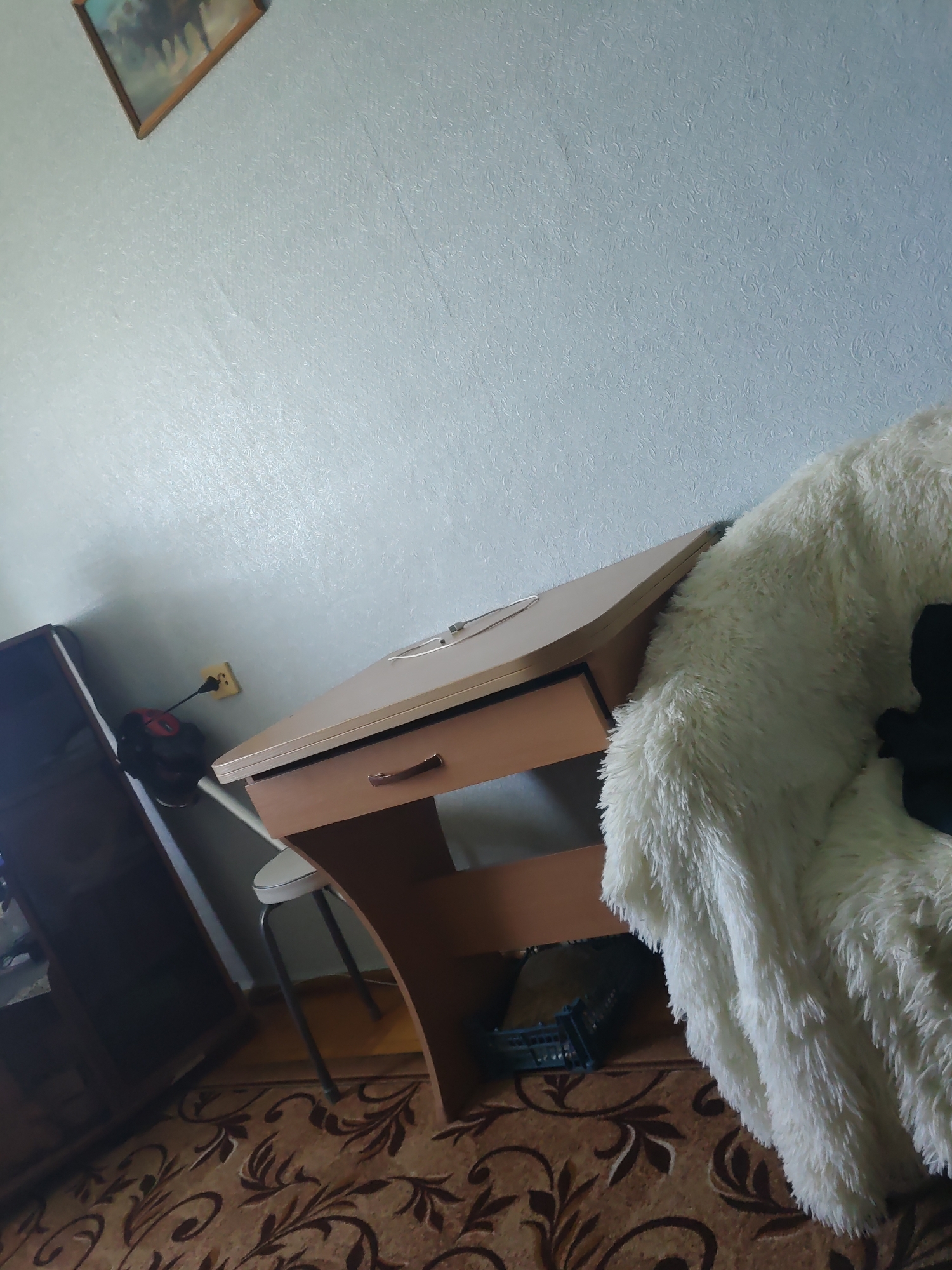 The image depicts an indoor scene, likely a room in a dwelling in Курашим, Russia. In the foreground, a portion of a white, fluffy blanket is visible, draped over an unseen object, possibly a chair or sofa. To the left of the blanket is a light brown wooden desk with a single drawer. A thin, white stool is positioned beneath the desk. On the wall behind the desk, a yellow electrical outlet is mounted, with a black and red fan plugged into it. To the far left of the image, a dark brown cabinet or wardrobe is partially visible. The floor is covered by a patterned rug in shades of brown and beige. A framed picture, featuring what appears to be horses, hangs on the light blue, textured wall in the upper left corner. The lighting suggests it is daytime.