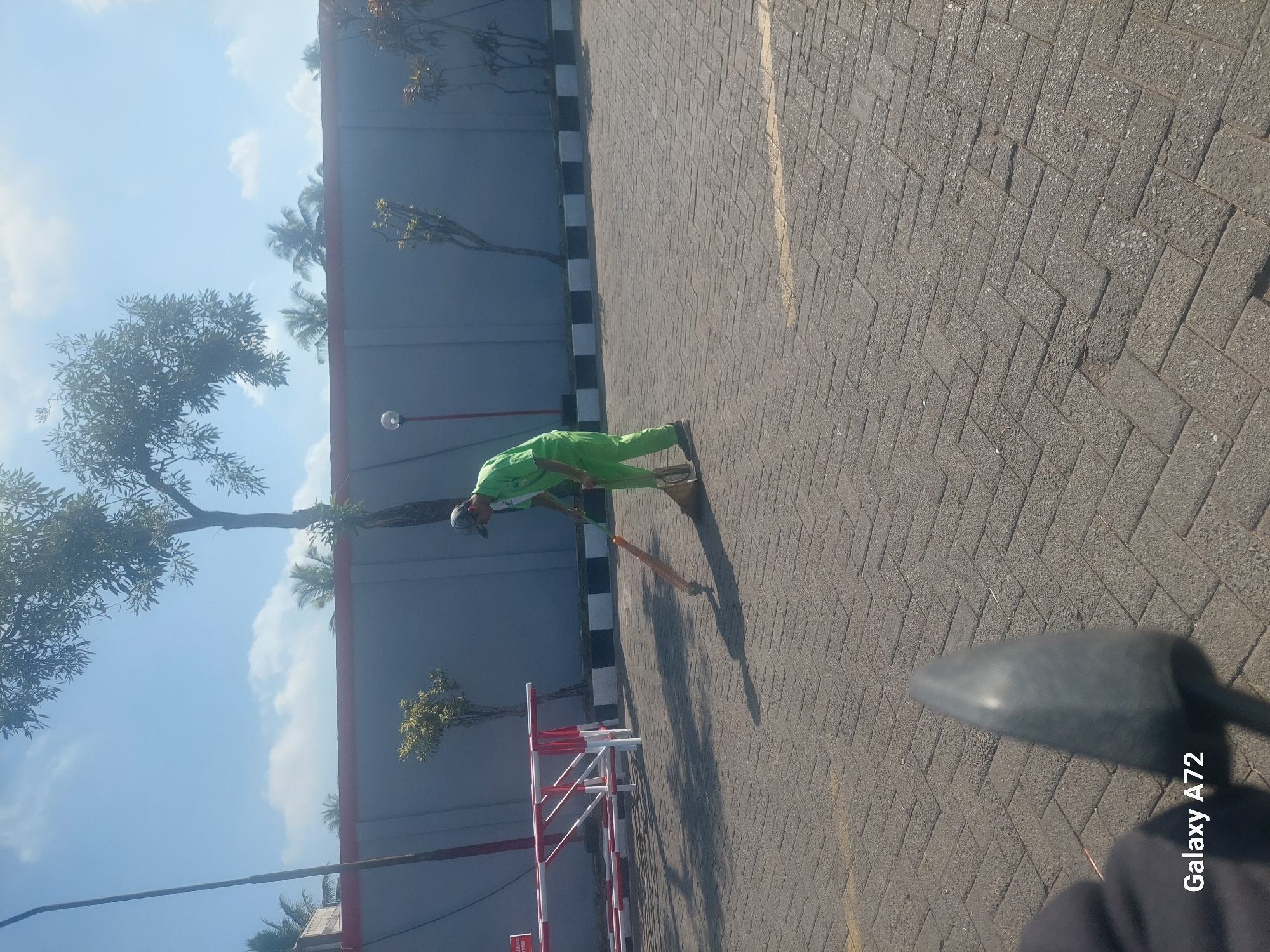 A person wearing a green uniform and a baseball cap is sweeping a paved area with a broom. The person is positioned in the middle of the frame, facing towards the right. The ground is paved with interlocking bricks, and a yellow line is visible on the ground in front of the person. A red and white striped barrier is visible in the background to the left, and a gray wall runs along the back of the scene. Trees and palm trees are visible beyond the wall. The sky is blue with some clouds. The image appears to have been taken outdoors in a city. The text "Galaxy A72" is visible in the bottom left corner.