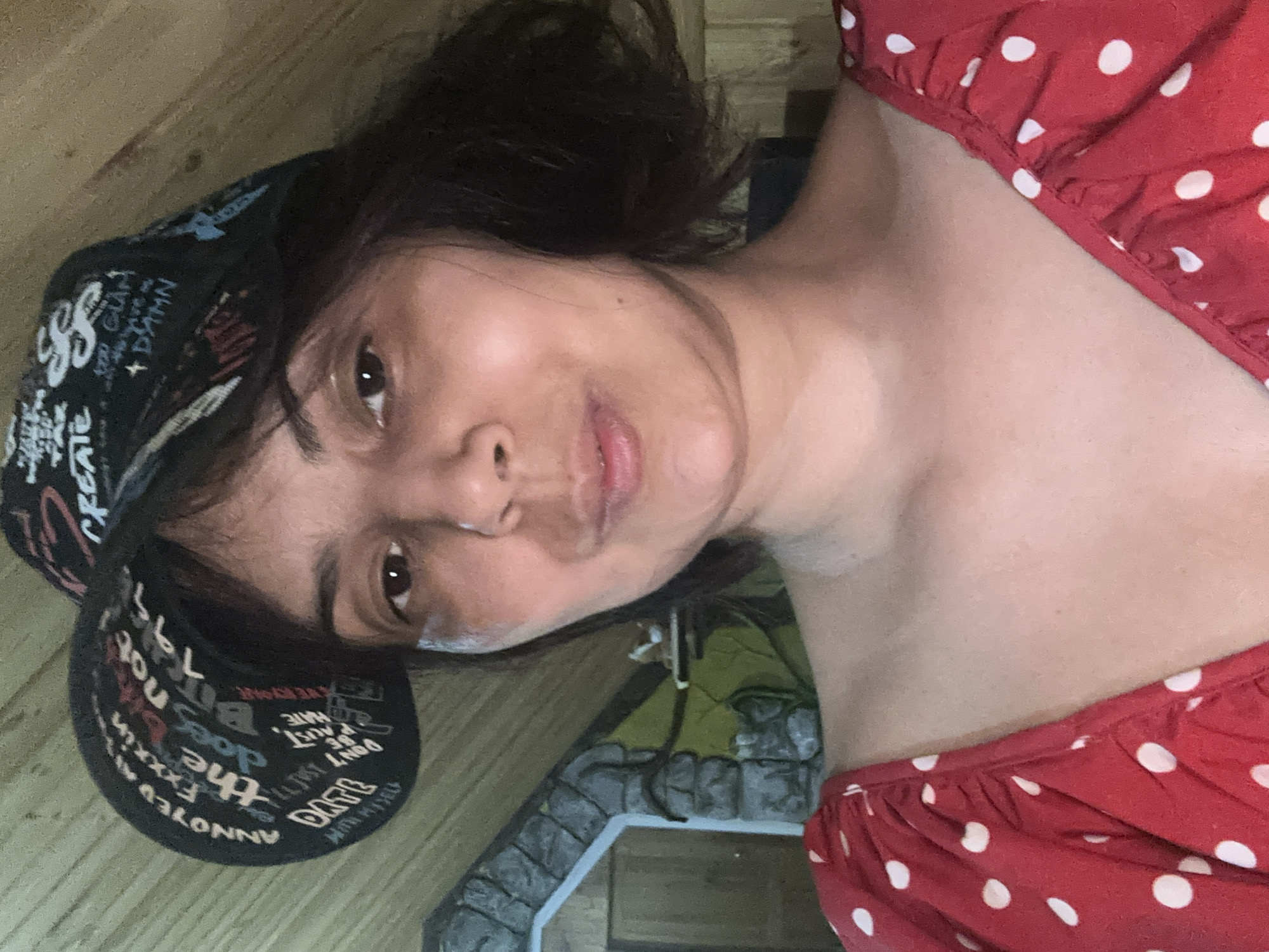 A woman is shown in a close-up, selfie-style shot. She is wearing a black baseball cap with white lettering, and a red dress with white polka dots. The background consists of a wooden ceiling and a stone-like structure. The lighting is warm and appears to be indoors. The specific location in Kalininskaya, Russia is not discernible from the image.