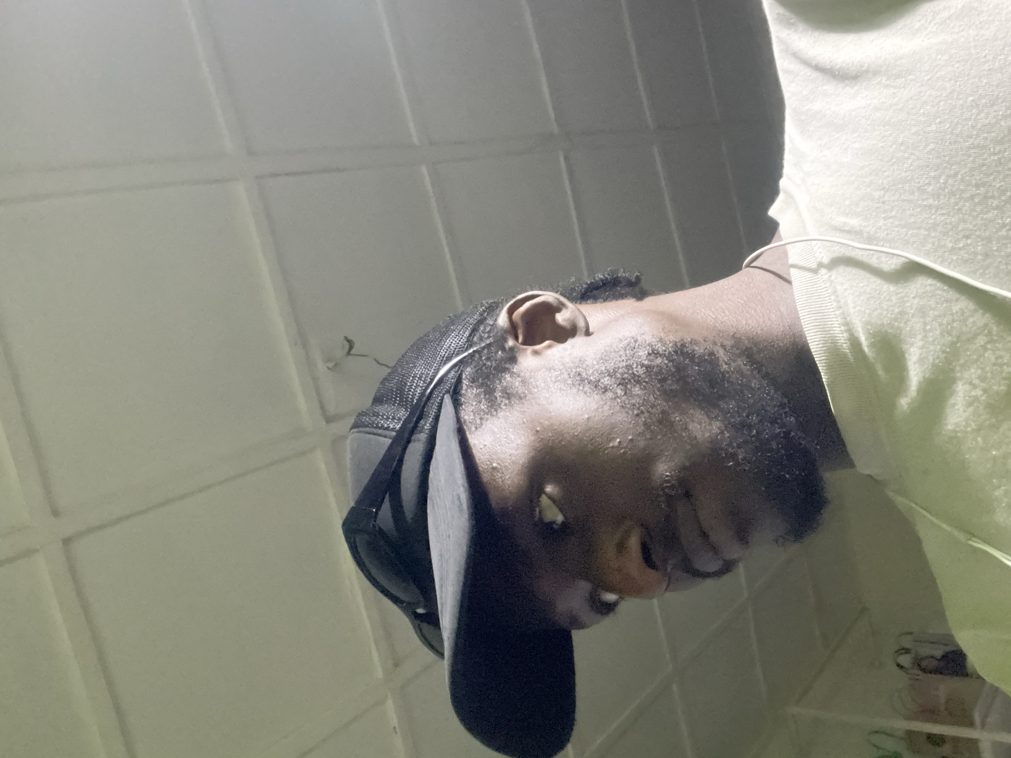 The image is a medium shot of a Black male wearing a black baseball cap with a mesh back and sunglasses resting on top. He is looking off to his left, and his face is partially obscured by the cap's brim. He has a light beard and is wearing a light green t-shirt. A white wire, likely from earphones, hangs down his chest. The background appears to be a light-colored ceiling with a grid pattern. The overall lighting is dim, with some light source casting shadows. There is no discernible indication of the image being taken in Jos, Nigeria.