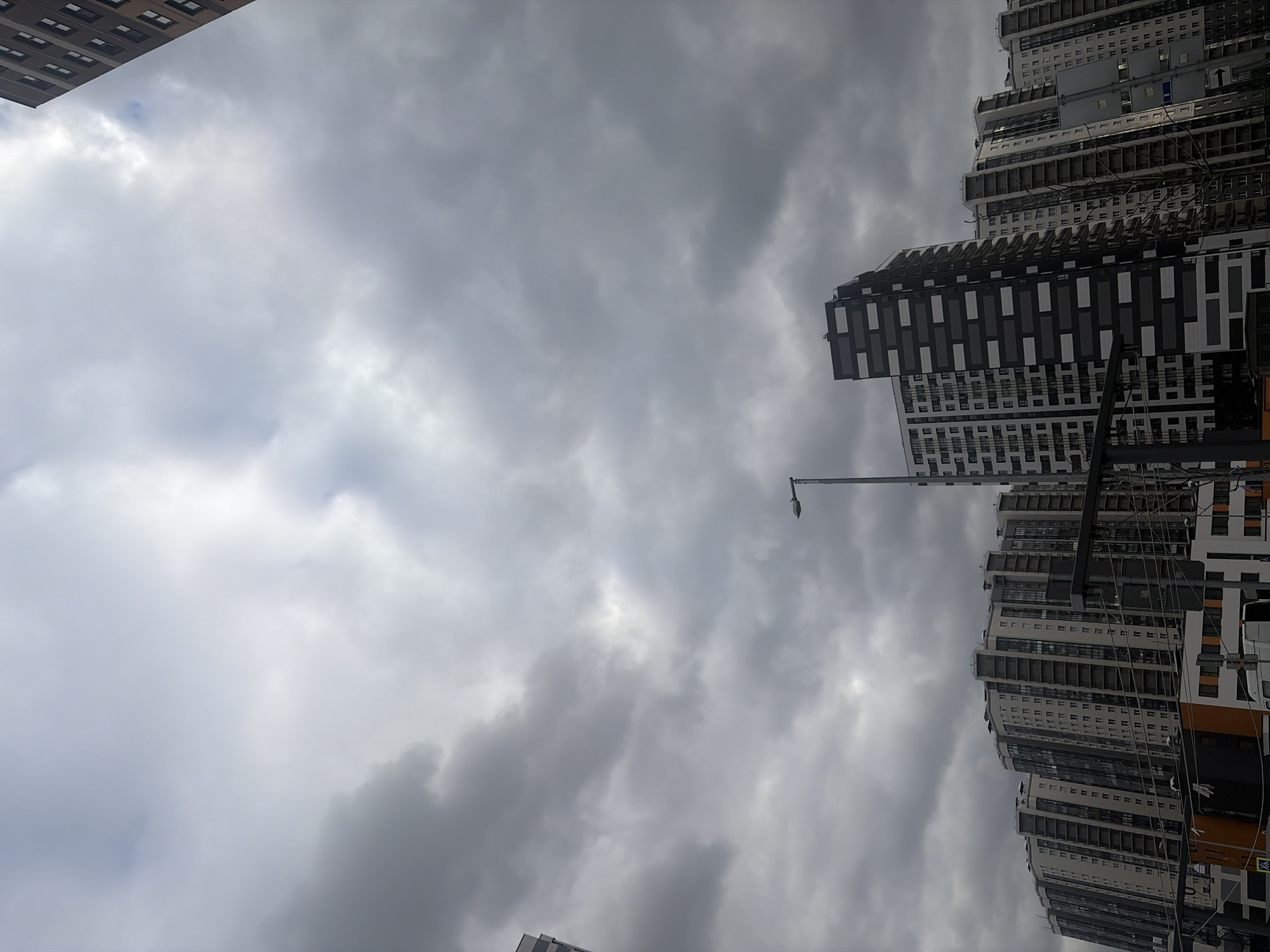 The image displays an urban landscape under a heavily overcast sky, likely in Saint Petersburg, Russia. The sky, filled with dense grey and white clouds, dominates the upper and central portions of the frame, indicating a dull or potentially rainy day with no direct sunlight.
Tall, modern multi-story buildings are prominent features of the scene. On the far left, the corner of a building with dark-framed rectangular windows is visible at the top. The right side of the image is largely occupied by a complex of high-rise apartment buildings. These structures showcase contemporary architecture with numerous balconies and windows, featuring facades that mix white, dark grey, and black panels, some arranged in distinct rectangular patterns.
A slender, tall street lamppost extends from the lower central area upwards, reaching towards the middle of the cloudy sky. A dense network of dark utility cables or wires crisscrosses between the lamppost and the buildings on the right, suggesting a developed urban infrastructure. No people or specific activities are discernible, and there is no visible text within the image. The overall impression is of a bustling modern city on a subdued, cloudy day.