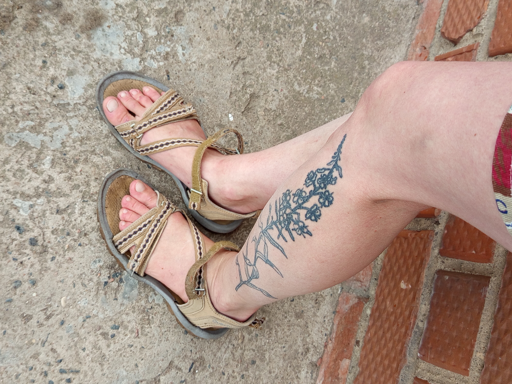 A person's legs, adorned with a tattoo of a sprig of flowers and grass on the left calf, are visible, wearing tan-colored sandals with crisscrossed straps. The individual is standing on a concrete surface, with a brick wall forming the background. The scene is set in Gdov, Russia.