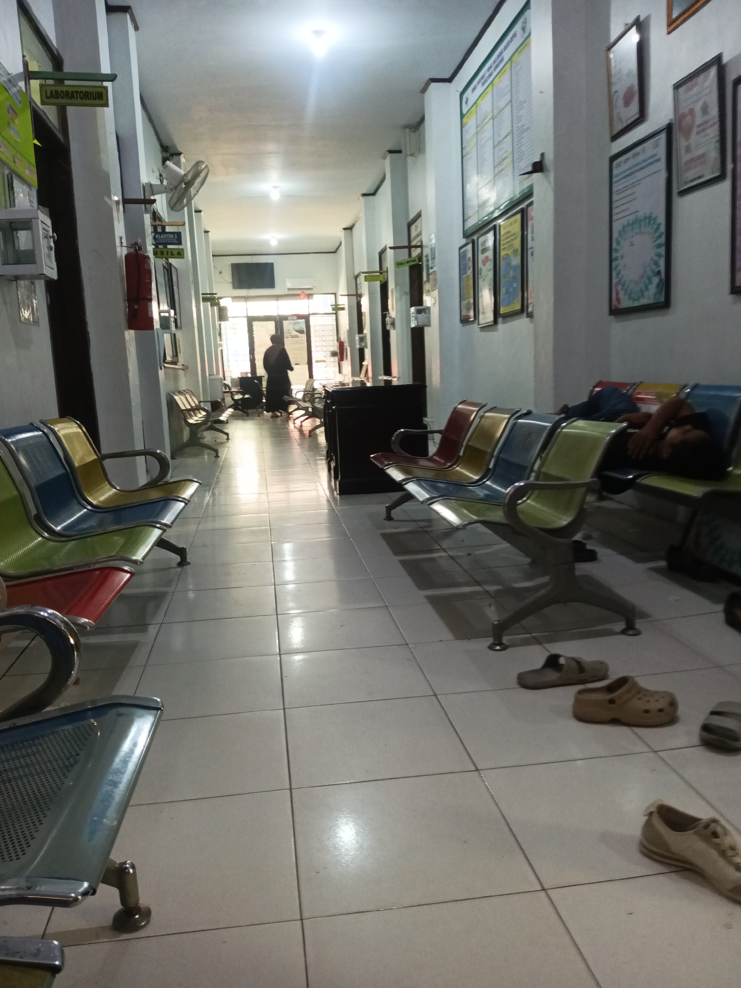 The image shows an indoor hallway at a medical facility, likely in Paddoang Doangan, Indonesia, with tiled flooring and white walls. Along the hallway are several rows of colorful metal chairs, some occupied and some empty. In the foreground, a person is lying down on a row of blue and green chairs, asleep. Further down the hall, a person in dark clothing is standing near a set of glass doors. Various signs and posters are mounted on the walls, including one labeled "LABORATORIUM". A fire extinguisher is visible on the left side of the hallway. On the floor, several pairs of footwear are scattered, including sandals and sneakers. The lighting appears to be fluorescent, with some reflections on the polished floor.