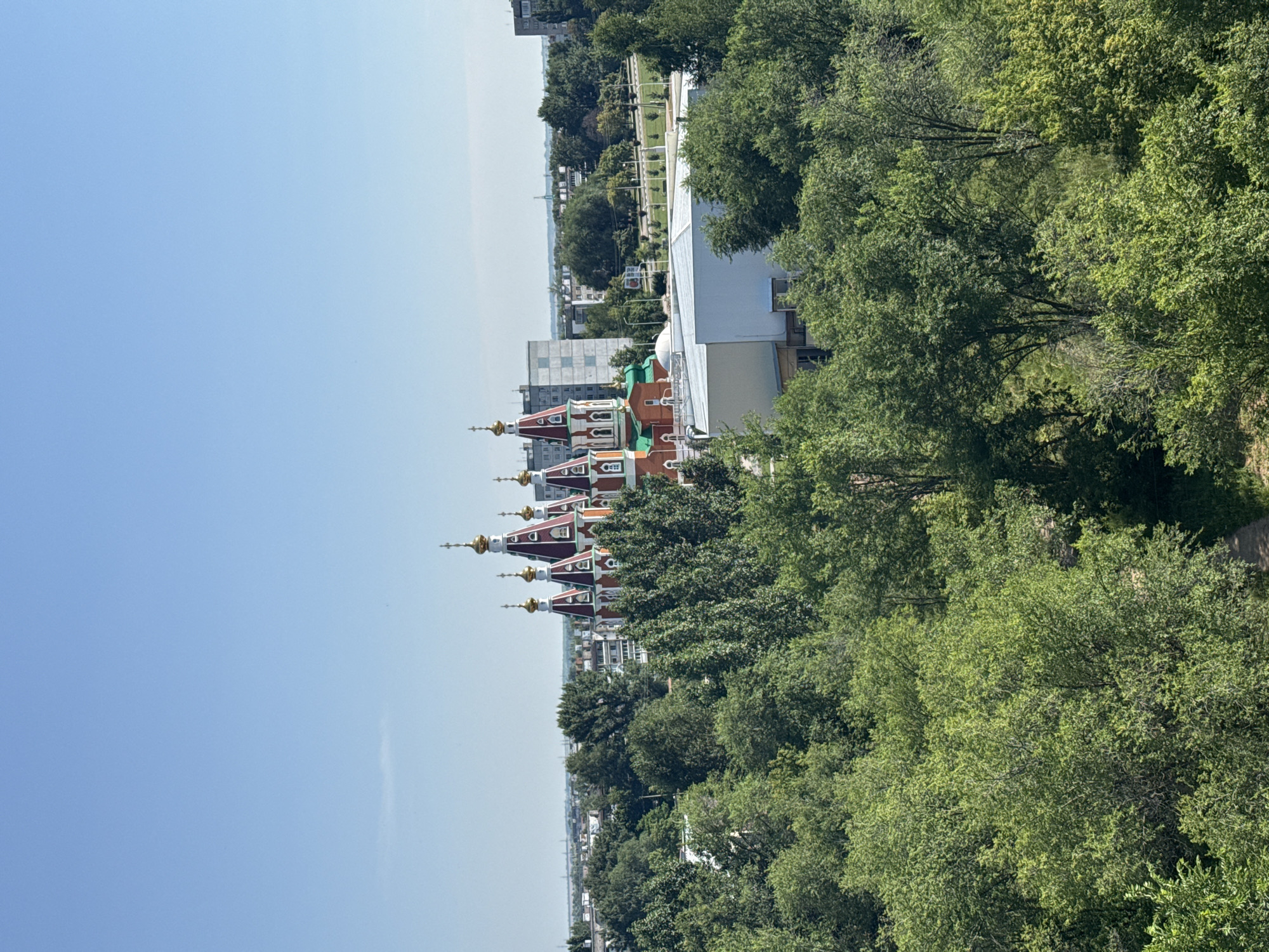 This is an aerial photograph, taken on a clear, sunny day, overlooking a cityscape dominated by lush green trees in the foreground. In the mid-ground, a distinctive red and white domed building, likely a church or cathedral with golden spires, stands prominently amidst other buildings and more trees. The background shows a flat urban landscape with a mix of residential and industrial buildings, stretching towards the horizon under a pale blue sky. The scene appears to be of the city of Otradny, Russia, given the architectural style of the religious building and the general urban setting. There are no people or specific actions depicted; the image is a static landscape view.