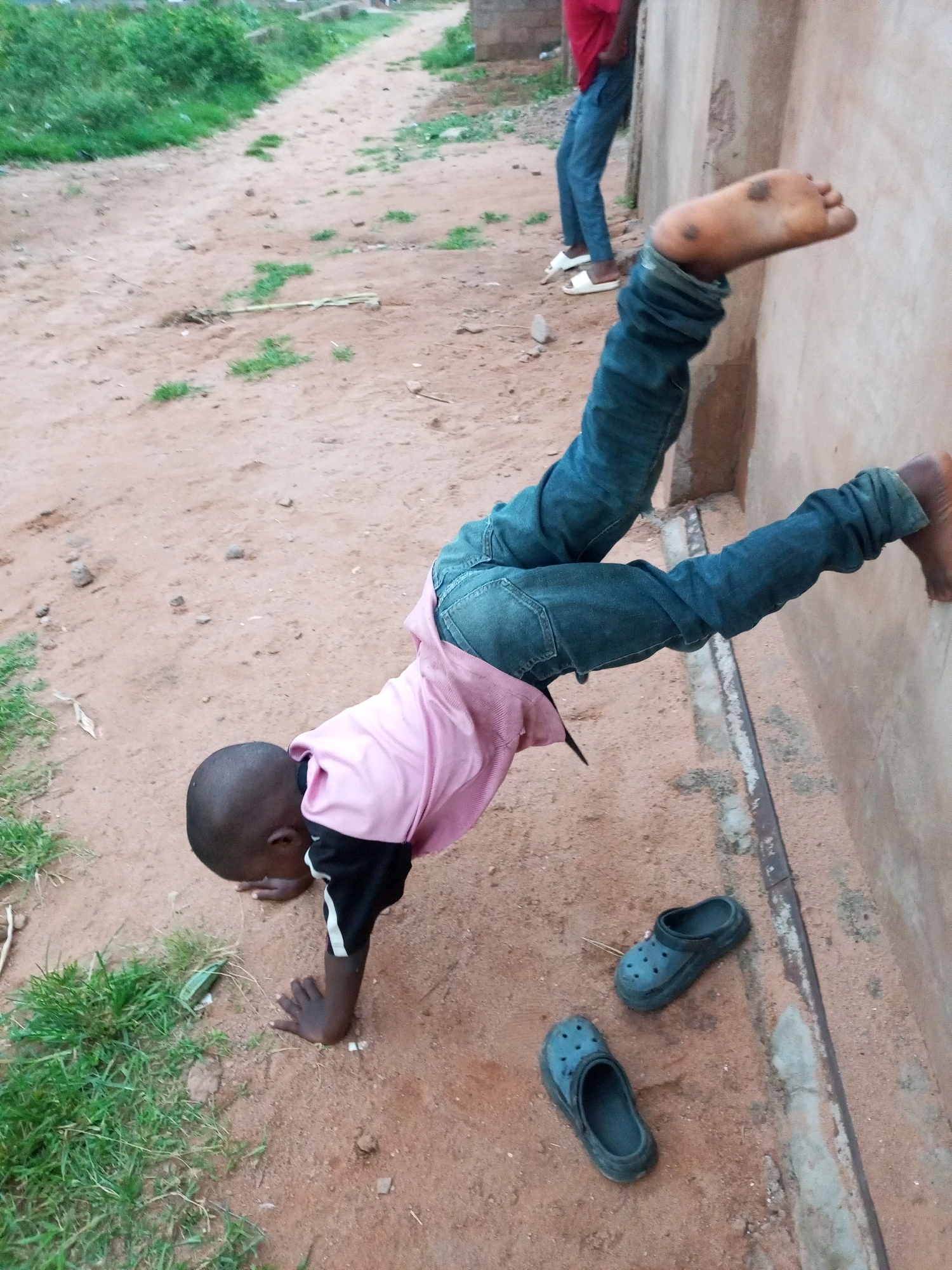 In the city of Tempure, Nigeria, a young child is seen performing a handstand against a building wall. The child is wearing a pink t-shirt with black and white accents on the sleeves and blue denim jeans. Their left hand is on the ground for support, and their legs are extended upwards, with their right foot resting against the wall. Two blue Crocs are lying on the sandy ground near the child. In the background, another individual in a red shirt and blue jeans stands near the wall, and a path lined with sparse vegetation is visible. The ground is covered in reddish-brown dirt and small stones.