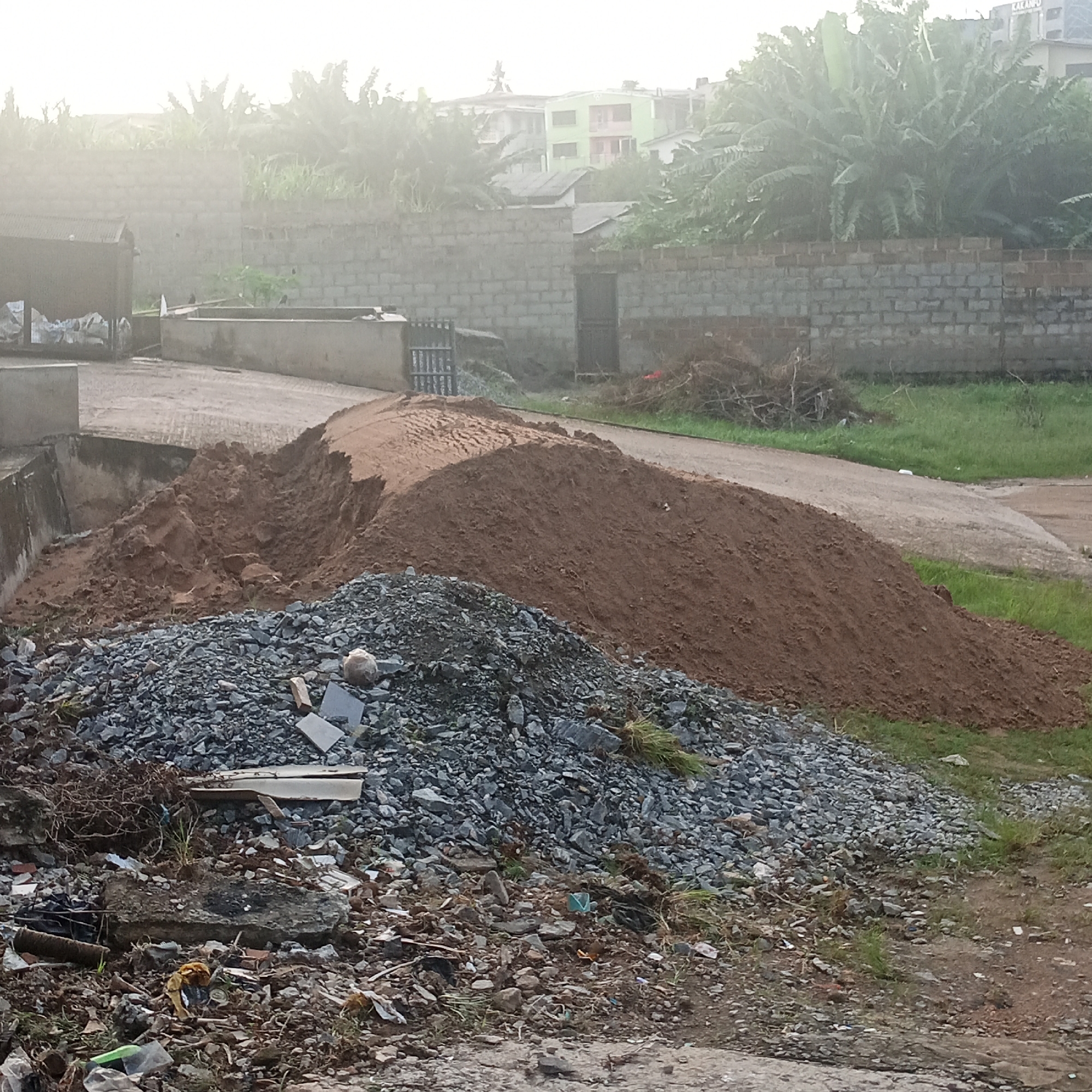 The image depicts an outdoor scene in an urban environment, likely in Ibadan, Nigeria. In the foreground, there are large piles of construction materials, including dark gray gravel and reddish-brown soil. Scattered amongst the gravel and soil are various debris items such as broken bricks, plastic fragments, and dried plant matter. Behind the piles of materials, a paved surface or road is visible, with patches of green grass along its edges. Further in the background, a brick wall runs horizontally across the image, separating the construction area from what appears to be a residential or built-up area. The background also features trees and buildings, with a particularly noticeable green building. The sky is overcast, suggesting a cloudy or hazy day. There are no people visible in the image. The scene appears to be a construction site or an area where construction materials are being stored or processed.