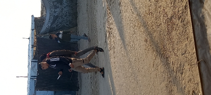 The scene is an unpaved, sandy expanse in Khan Yunis, Palestinian Territory, illuminated by a clear sky. Long shadows stretch across the ground from left to right. In the background, multiple makeshift structures are visible, constructed from corrugated metal sheets, blue and grey tarpaulins, and patterned fabric sheets. Three males are in motion, walking from left to right across the frame.
The male in the immediate foreground wears a dark jacket with three white stripes on the sleeves, light brown trousers with black side stripes, and black sandals. He holds a white-cased smartphone in his left hand and wears a watch on his left wrist, with his gaze directed downwards. Directly behind him, a second male is partially visible, dressed in dark clothing, walking in the same direction. To the right, a third, younger male wears a horizontally striped long-sleeved shirt and light blue jeans, with his right hand positioned at his mouth. The sandy ground displays multiple footprints and faint tire tracks.
