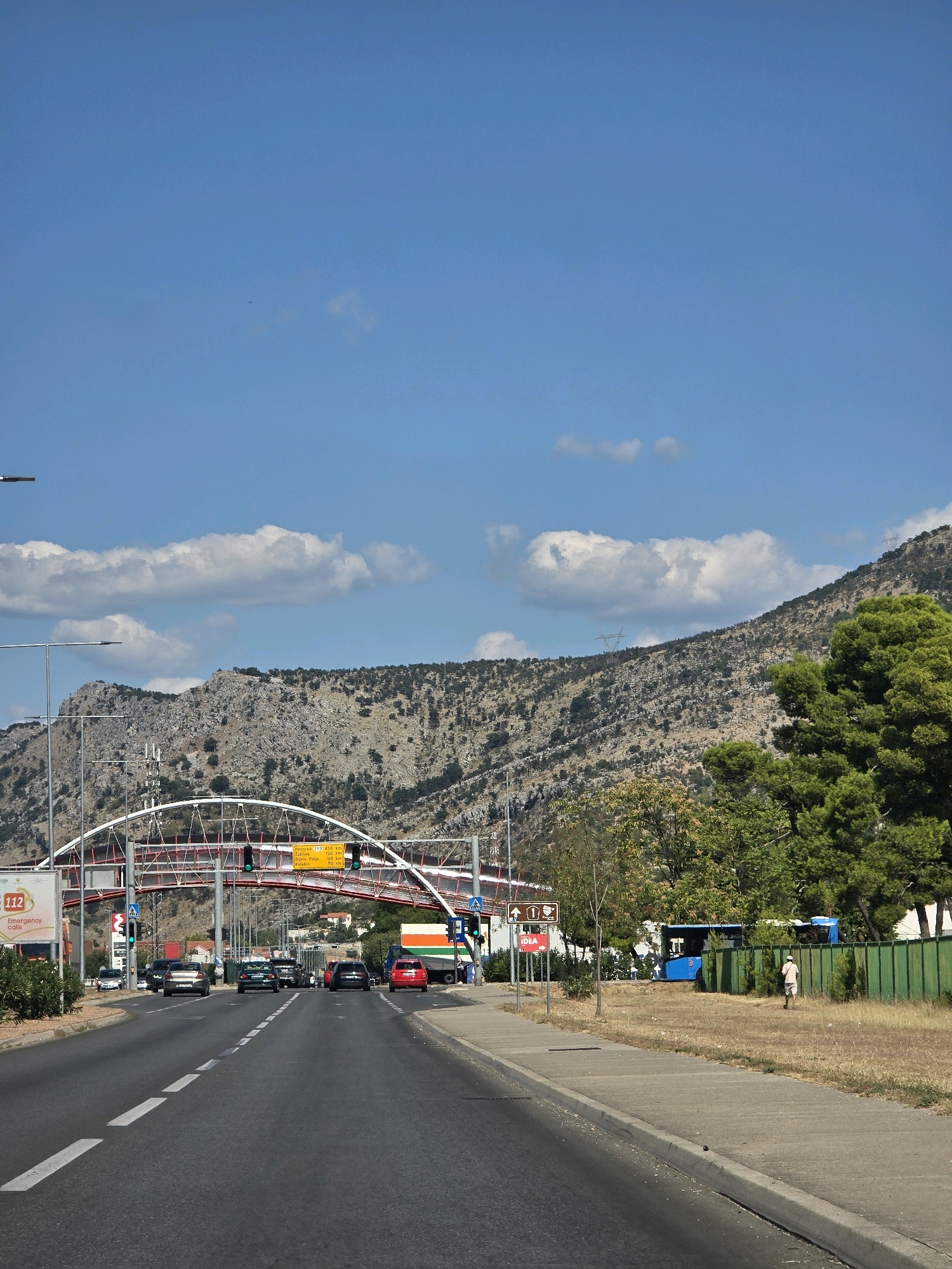 The image shows a street scene in Podgorica, Montenegro. A highway with multiple lanes runs through the foreground, with cars driving in both directions. A pedestrian bridge arches over the highway, with signs indicating distances to nearby cities. The surrounding environment includes a dry, grassy area with scattered trees and bushes, and a green fence running along the roadside. In the background, rocky hills rise, covered with sparse vegetation. The sky is clear blue with a few white clouds. A blue bus and a person in light clothing are visible on the right side of the road. A person wearing a white hat is seen walking on the right side of the image.