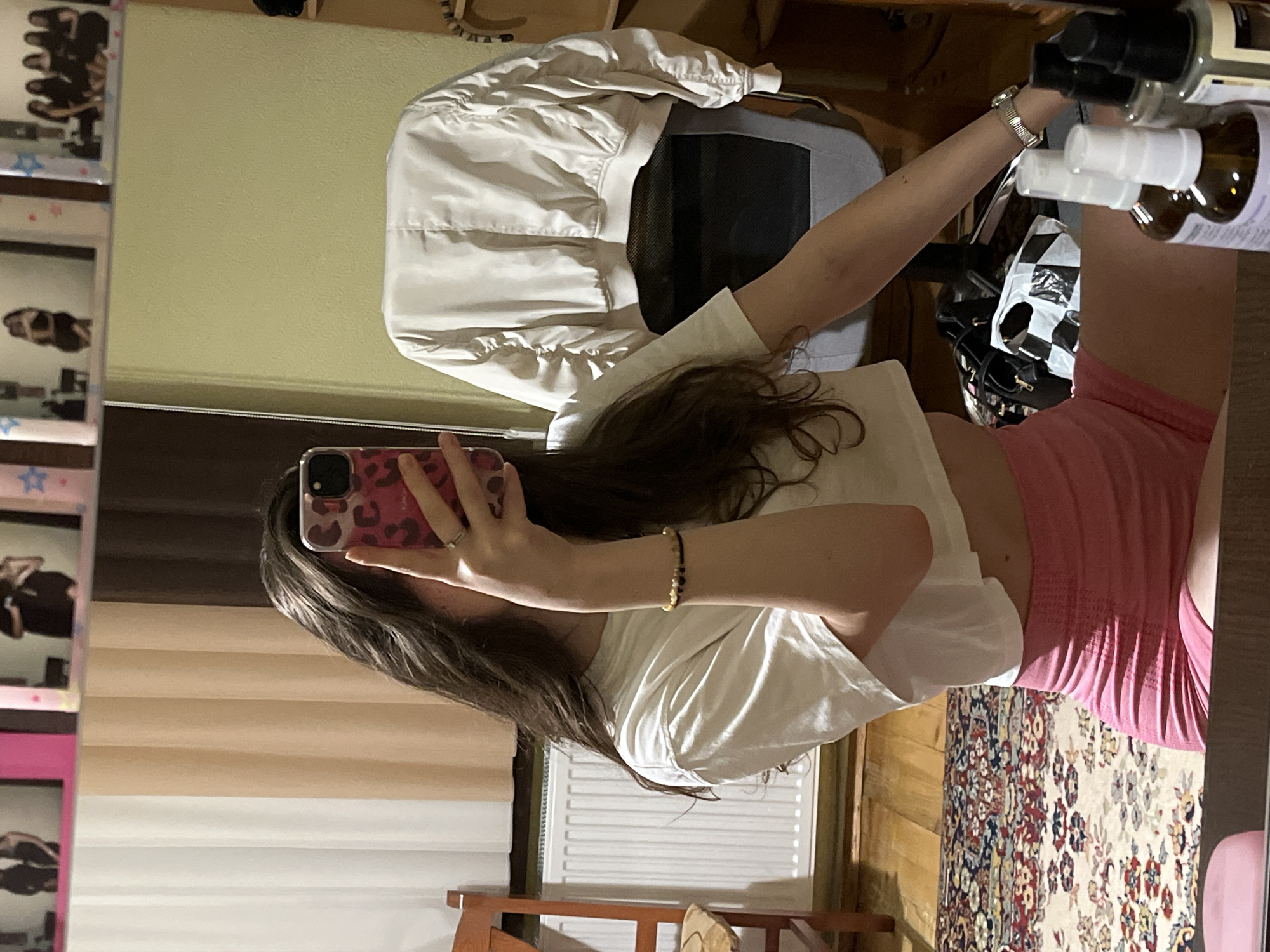 A female individual is depicted taking a mirror selfie within an indoor setting in Popintsi, Bulgaria. Her face is mostly obscured by a smartphone, which has a pink leopard print case and is held in her right hand. She possesses long, dark, wavy hair. She is attired in a white cropped t-shirt and pink form-fitting shorts. A beaded bracelet with dark and amber-colored beads is visible on her left wrist, and a silver ring is present on her right ring finger. A silver-toned watch is worn on her right wrist.
The reflection reveals a room with a light green wall to the right and white and beige vertically striped curtains to the left. A white radiator is positioned below the curtains. The flooring consists of wooden planks, partially covered by a patterned oriental-style rug. A light-colored bomber jacket is draped over the back of a grey office chair to the right. A wooden chair is partially visible on the far left. On the surface holding the mirror, several small bottles, some with spray tops, are arranged in the foreground. A black bag and a black-and-white patterned bag are visible on the floor behind these bottles. Above the main mirror reflection, multiple framed images or photos are visible, depicting individuals and groups of people. Some frames appear pink or have pink decorative elements, including small stars.