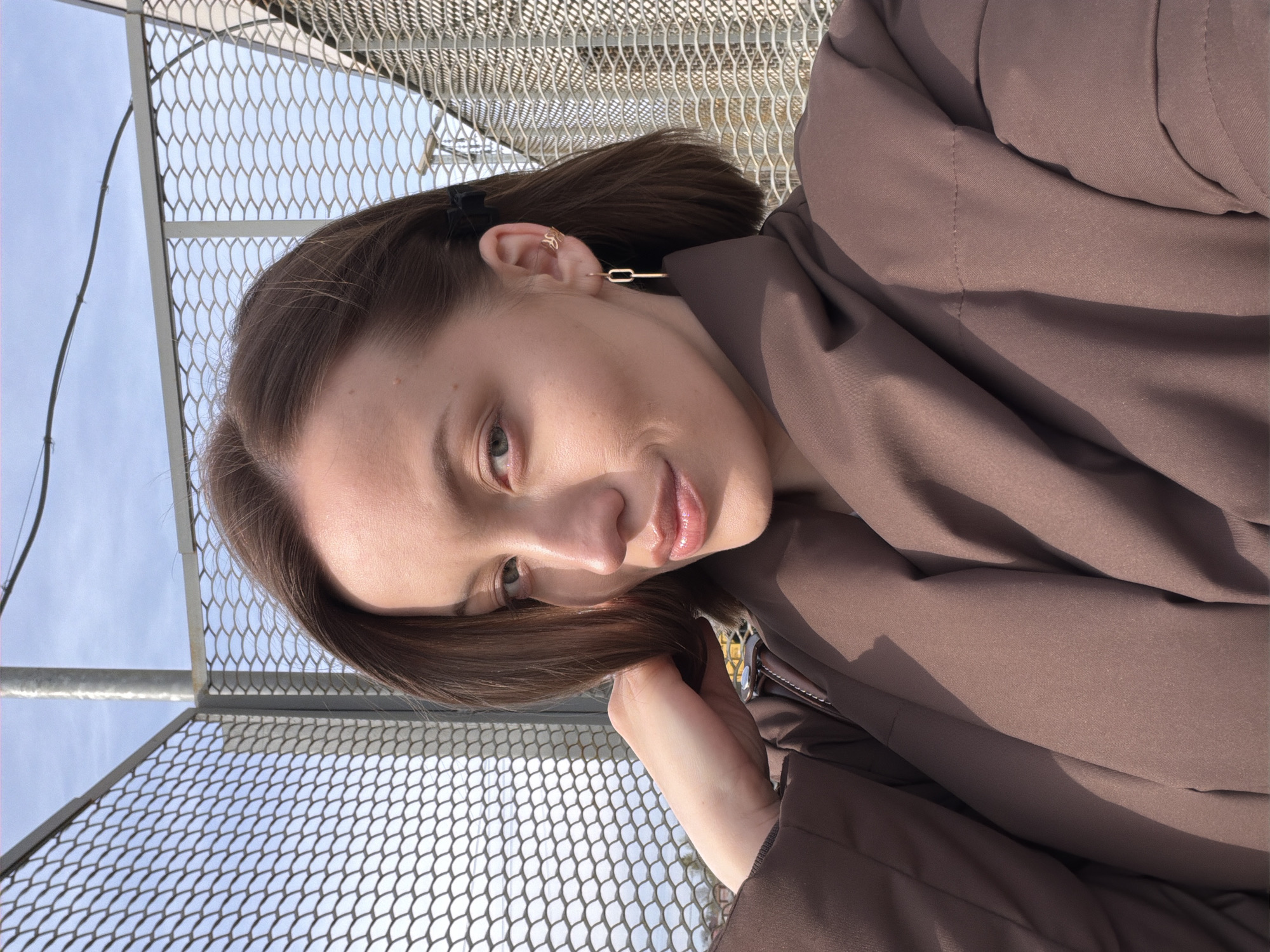A young woman with shoulder-length brown hair is pictured in front of a wire mesh fence in Reshetnikovo, Russia. She wears a brown jacket. A gold earring is visible in her left ear. The sky is visible through the mesh fence.