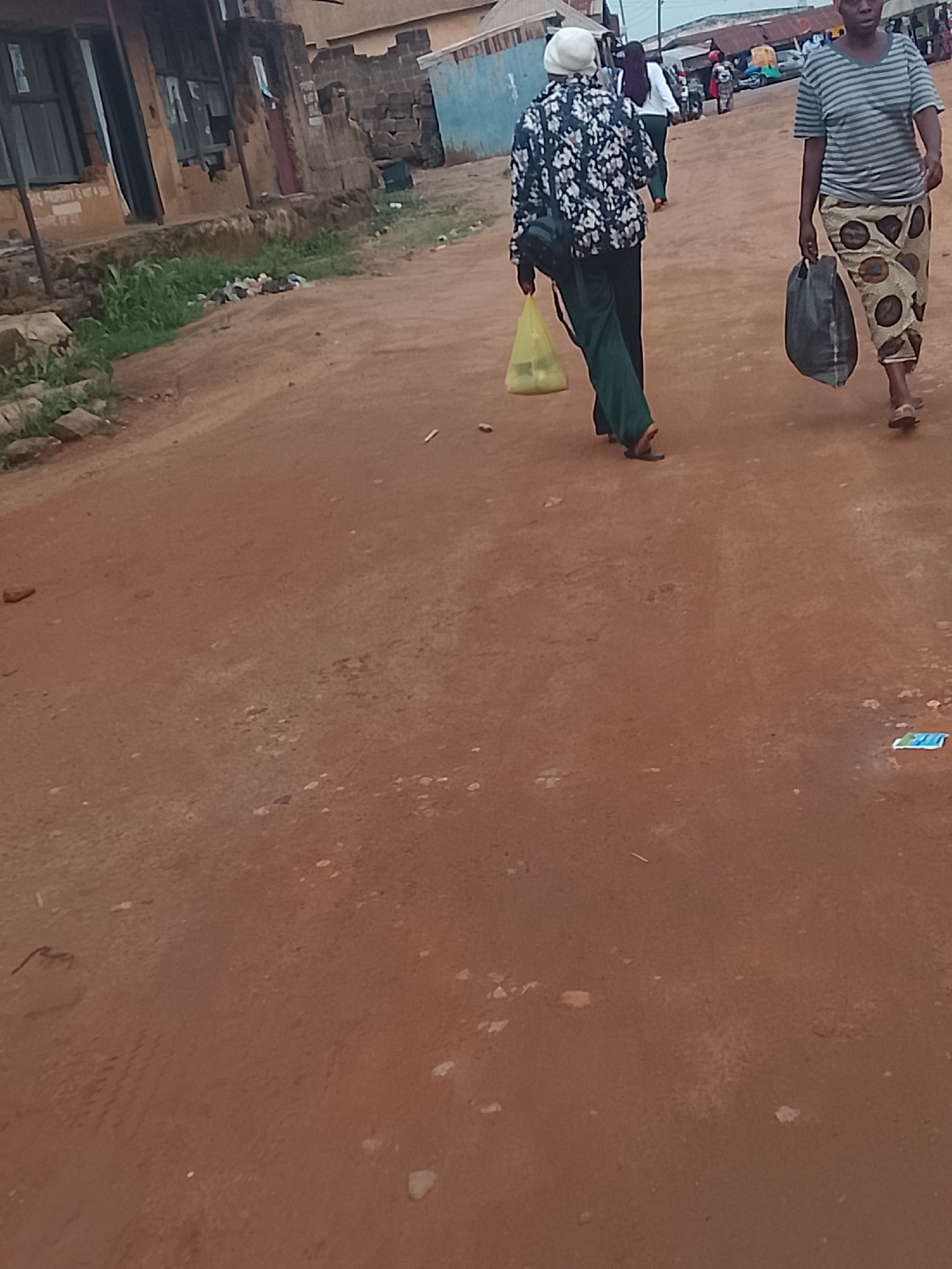 Two women walk away from the camera on a dirt road. The woman on the left wears a white head covering, a floral patterned jacket, dark green pants, and black sandals. She carries a yellow plastic bag. The woman on the right wears a striped shirt, a patterned skirt, and sandals, carrying a dark-colored bag. The dirt road is reddish-brown. Buildings are visible on the left side of the road, while other people and structures can be seen in the background. Based on visual characteristics, the image is likely taken in a rural area of Jos, Nigeria, though specific landmarks are not discernible.