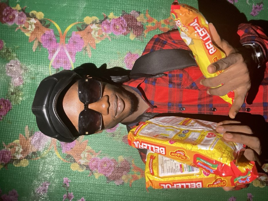 A man is lying on a surface with a green floral pattern, wearing a red plaid shirt, a black cap, and sunglasses. He holds three packages of "Belleful" chicken flavor noodles in his hands. He has a dark complexion and a small beard. The lighting in the photo is uneven, with some areas appearing darker due to shadows.