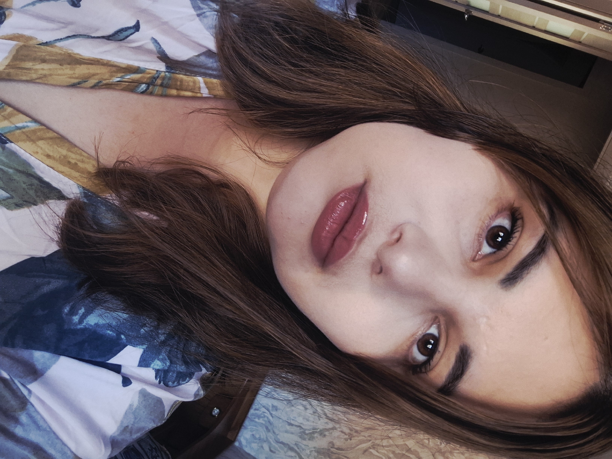 A close-up, angled shot of a woman's face, lying down. She has brown hair and light skin. Her lips are glossy, and she has brown eyes. The background consists of a patterned fabric, possibly a bedsheet, with a mix of blue, white, and yellow hues. There's also a dark, shadowed area in the upper right corner of the image, possibly indicating a wall or furniture. The location in the city of Амзя, Russia is unknown.