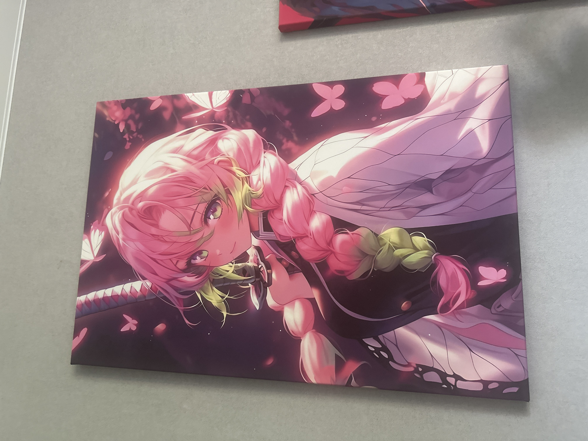 A framed anime-style artwork is displayed on a gray wall. The artwork features a young female character with long, braided pink hair and green eyes, dressed in a black and white outfit. She is holding a sword and surrounded by pink butterflies and floral elements. The artwork has a vibrant, colorful aesthetic.